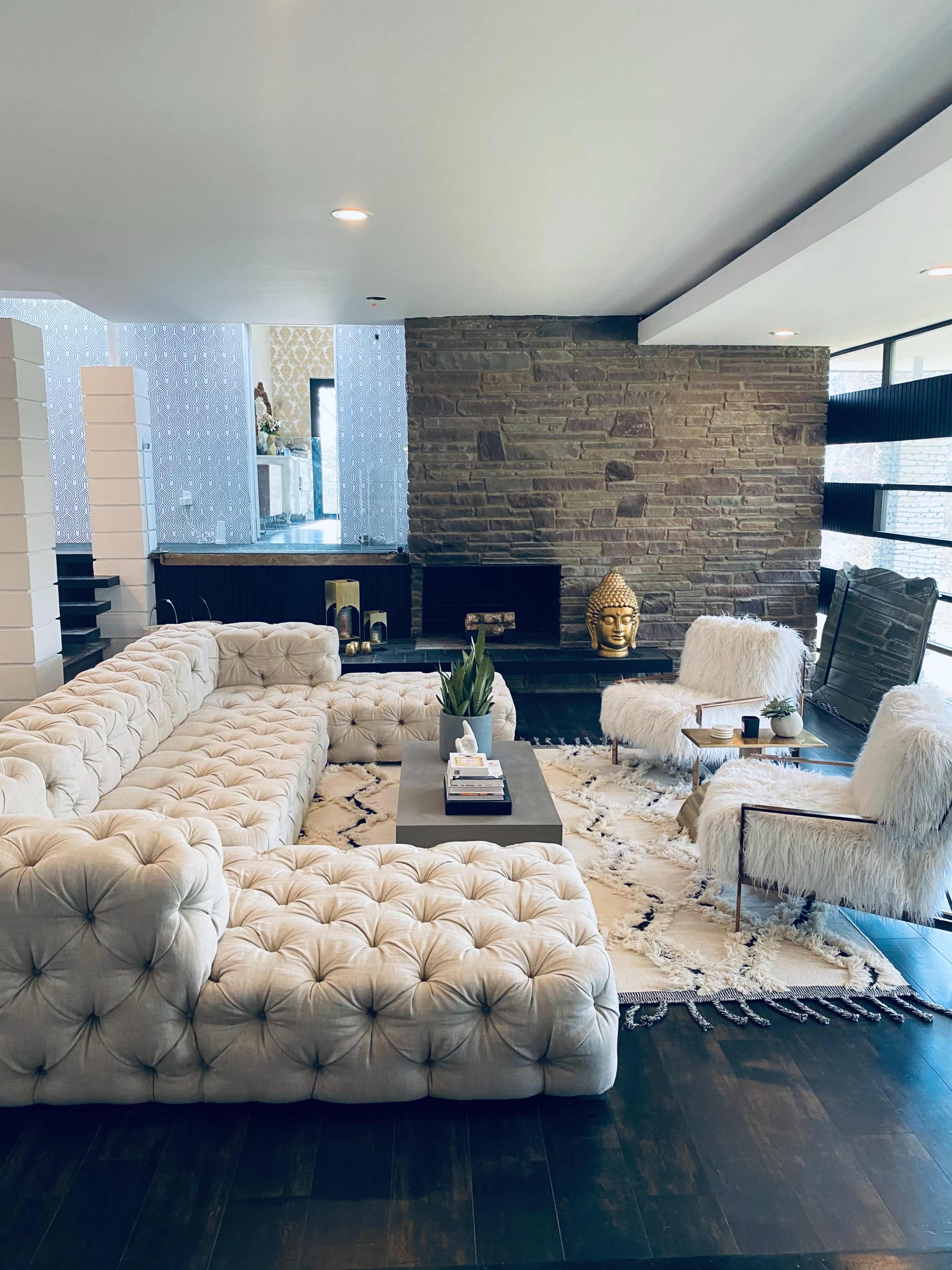 A modern living room featuring a large tufted sectional sofa, two accent chairs, a stone fireplace, and a decorative area rug.