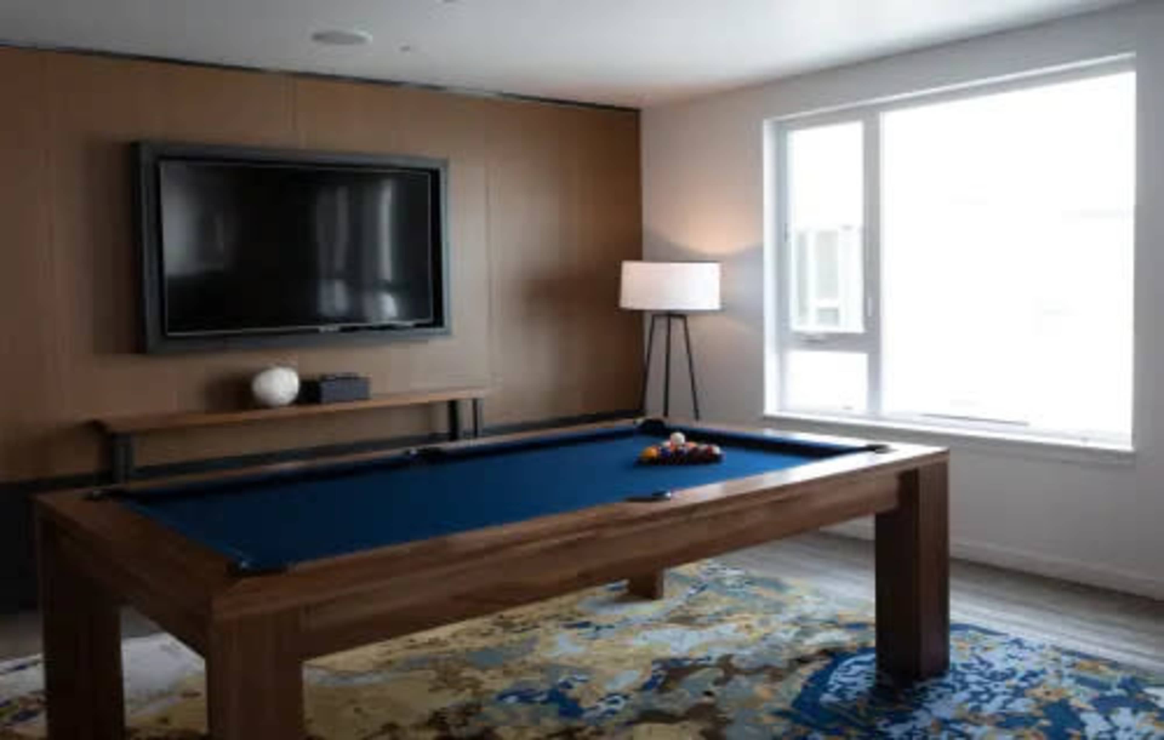 A modern game room featuring a pool table, a lamp, a television mounted on the wall, and a large window allowing natural light.