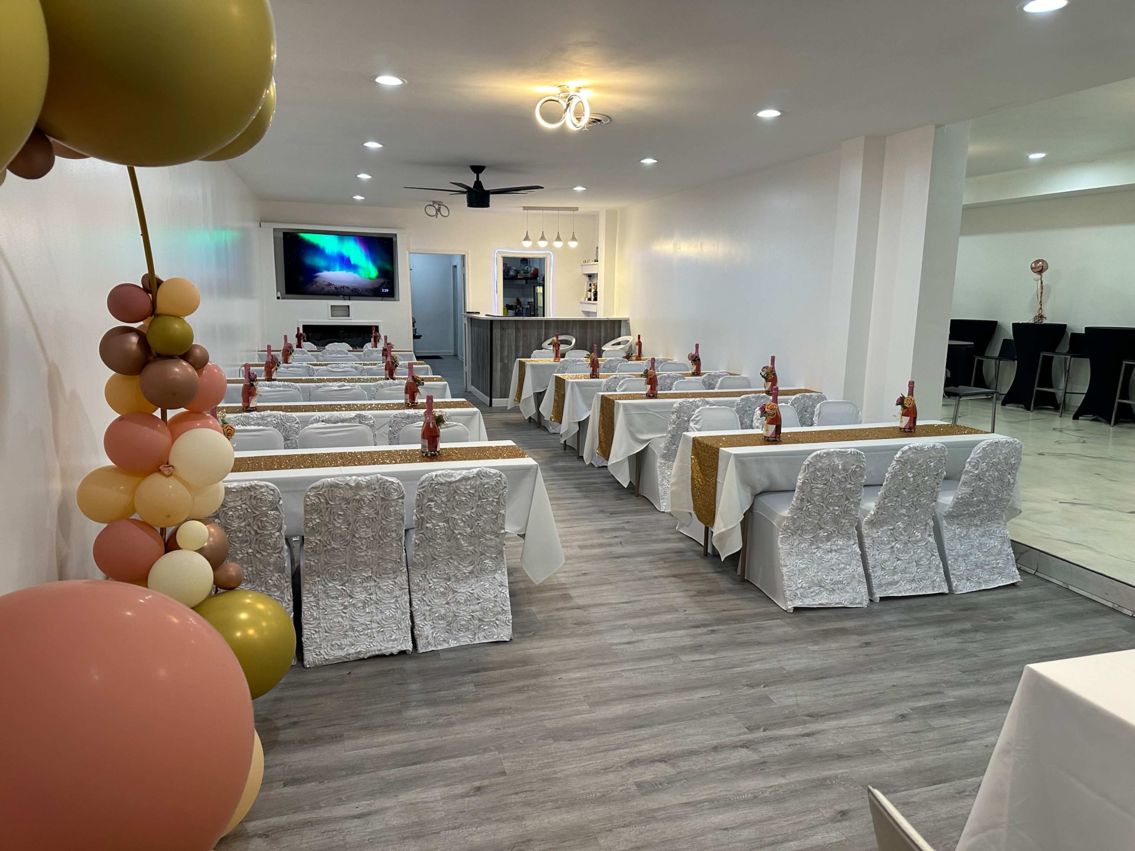 The image shows a banquet hall arranged with multiple tables set for an event, featuring decorative chairs and a backdrop with a television screen.