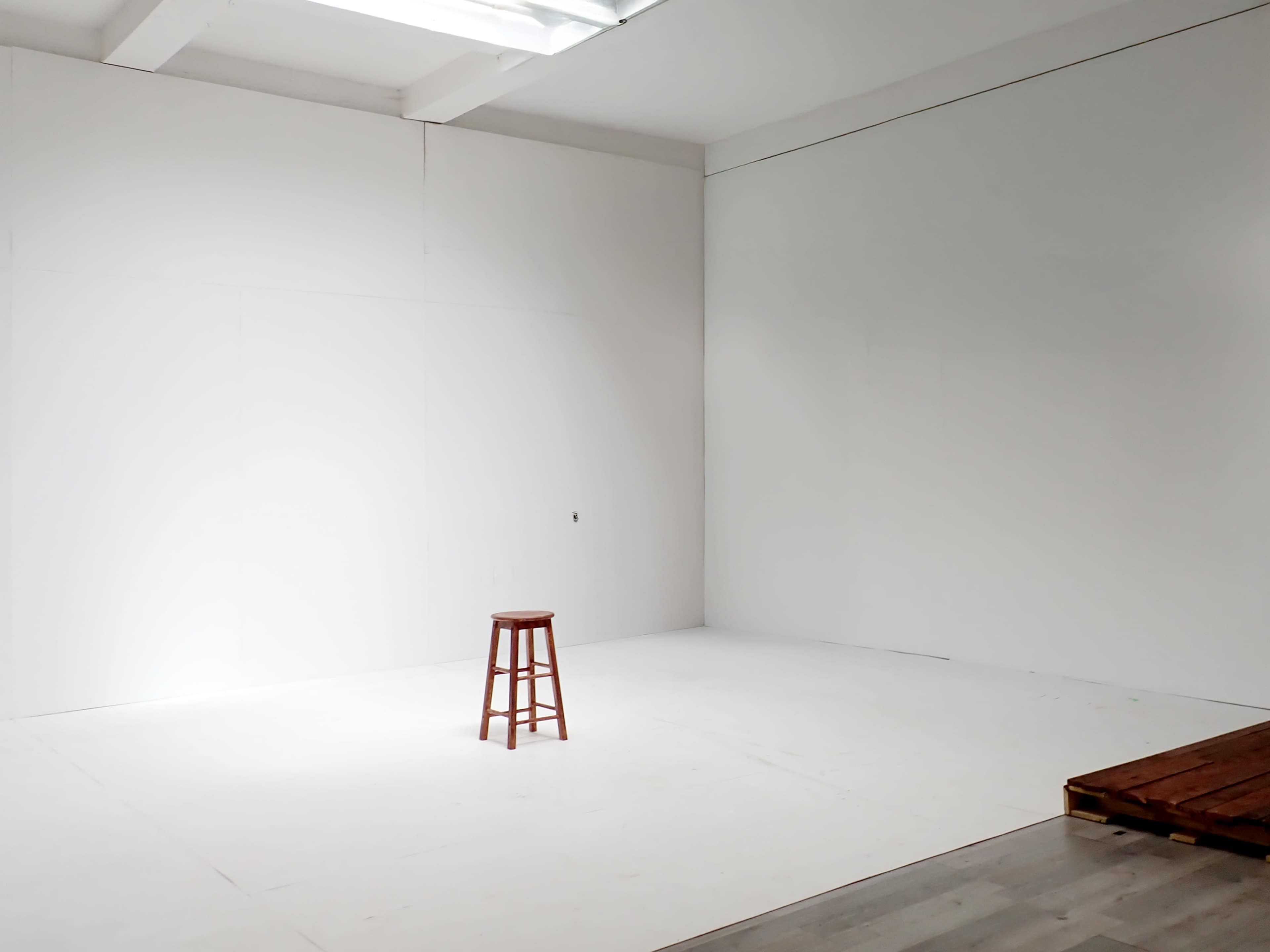 The image shows an empty room with white walls and a single wooden stool positioned in the center.