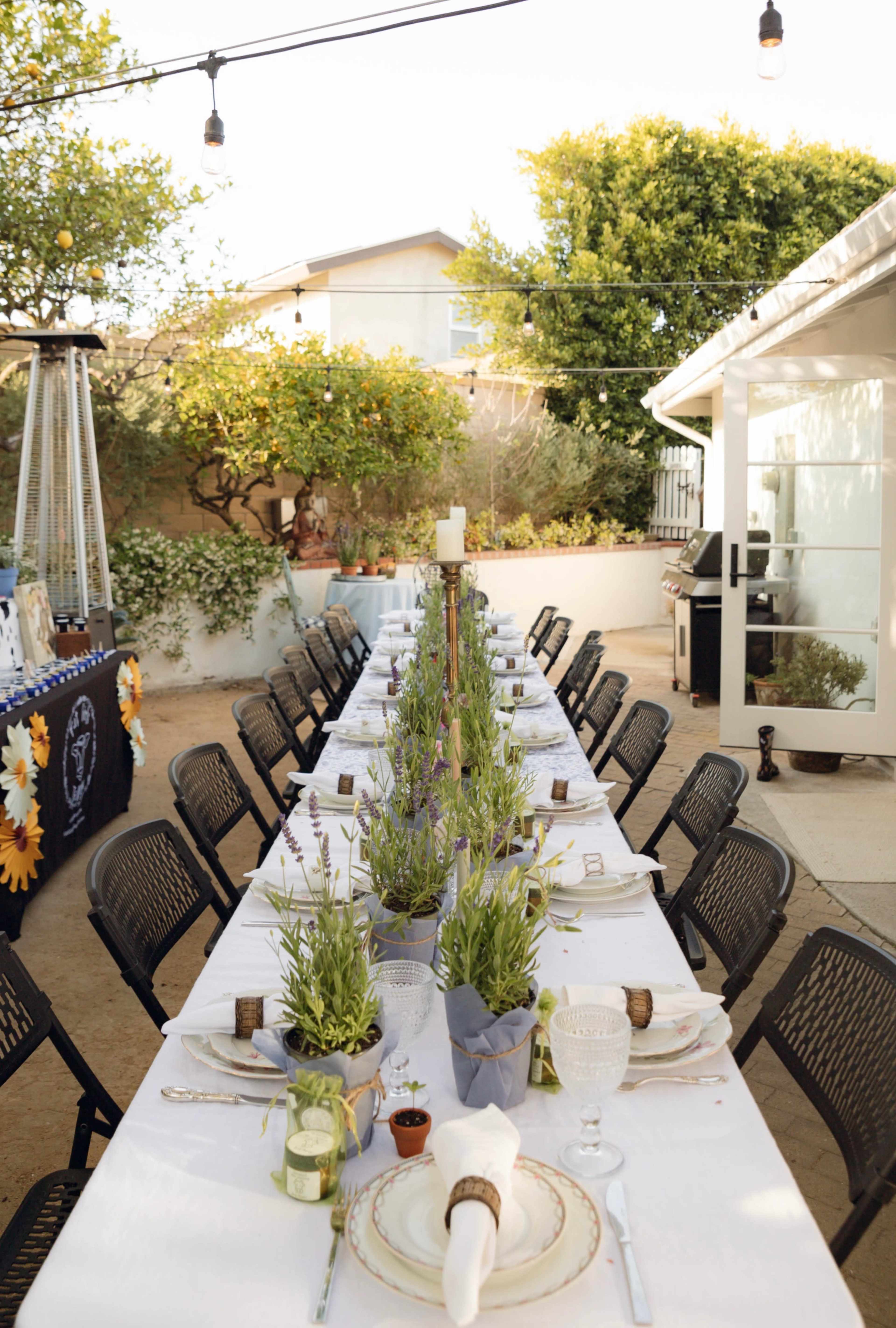 A long outdoor dining table is set with white tablecloths and potted herbs, surrounded by black folding chairs under string lights and trees.