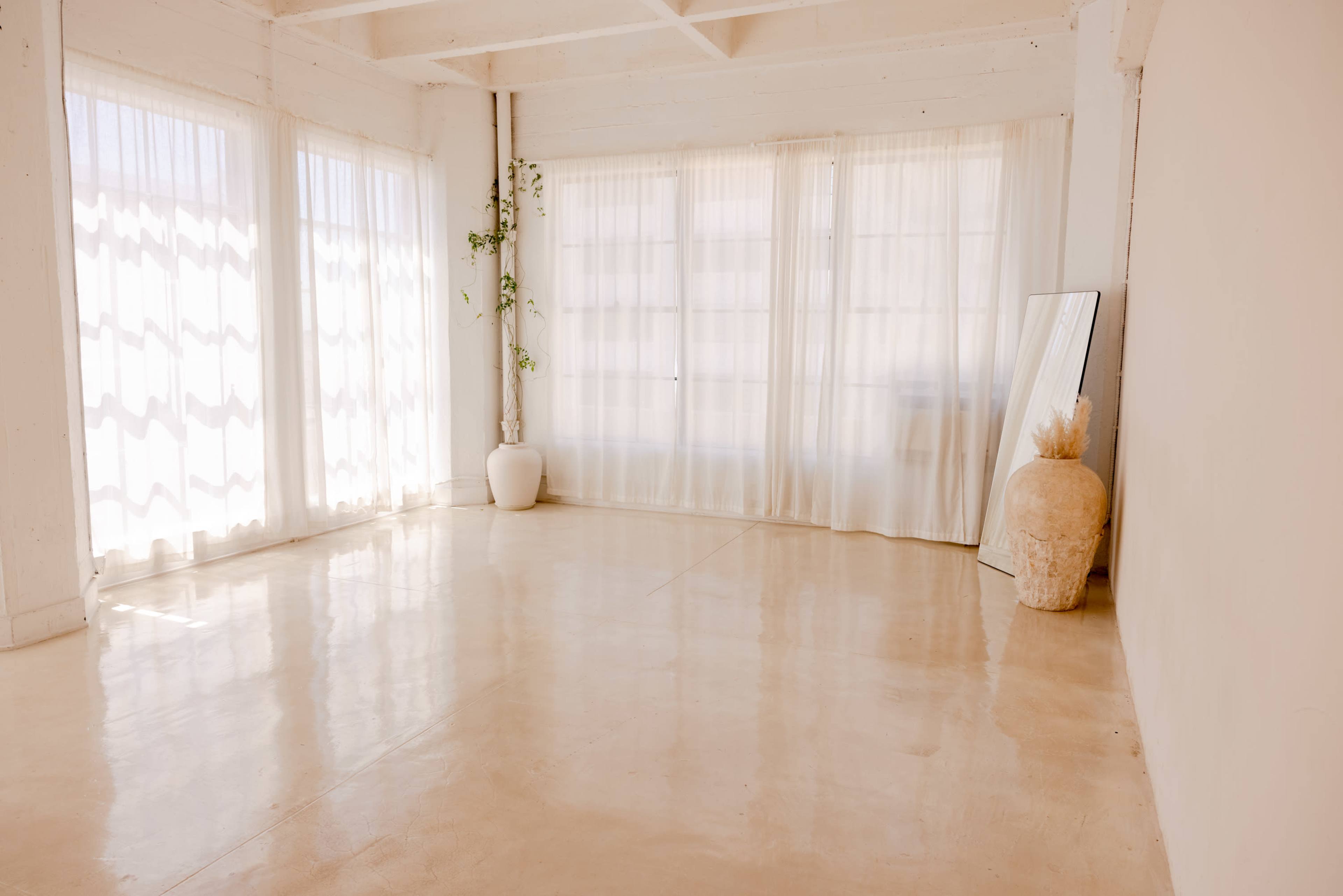 A bright, empty room with large windows covered by sheer curtains and a few decorative items, including a potted plant and a mirror.