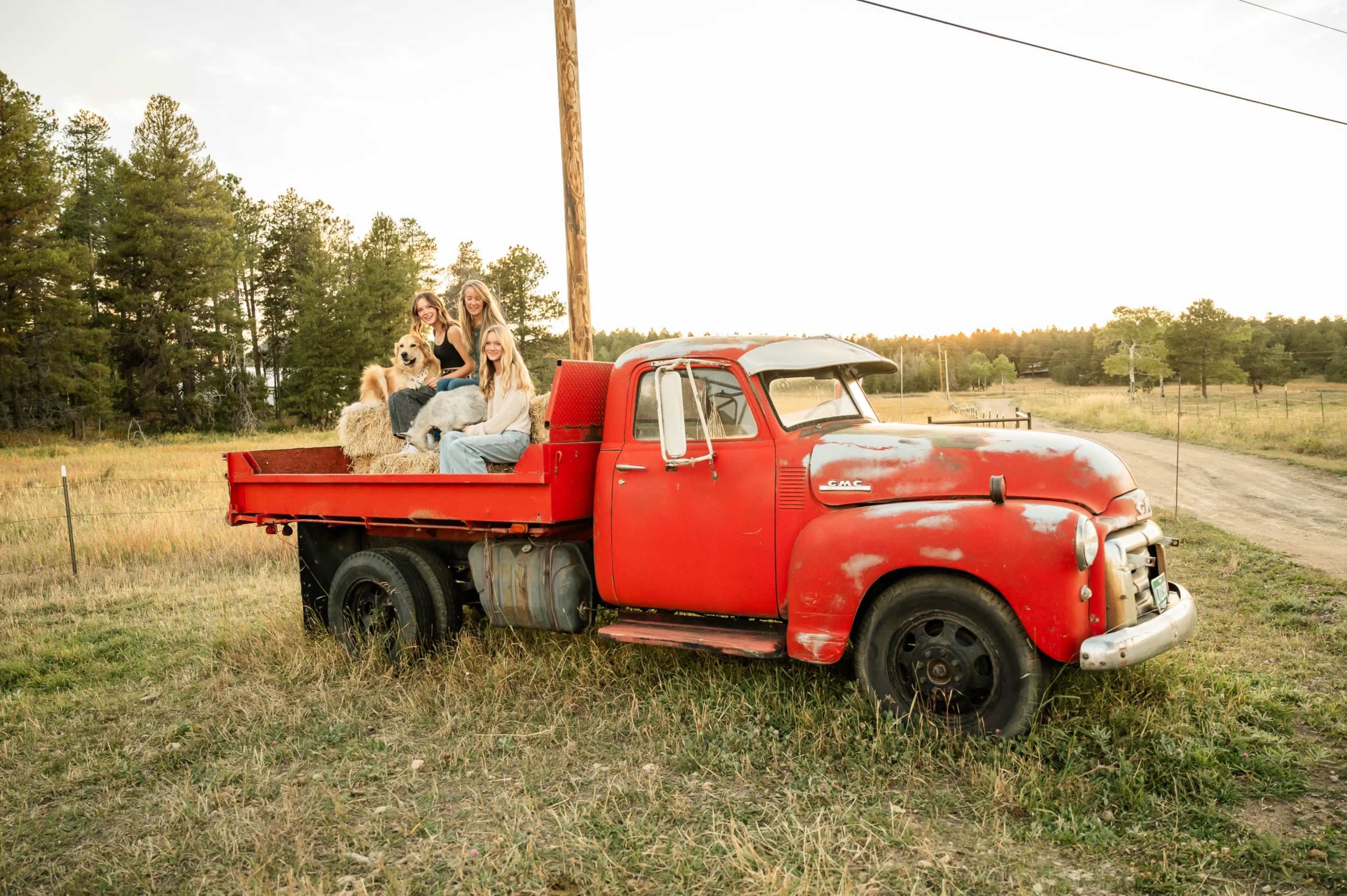 A group of four women and a dog are sitting on the back of an old red pickup truck parked in a grassy field.