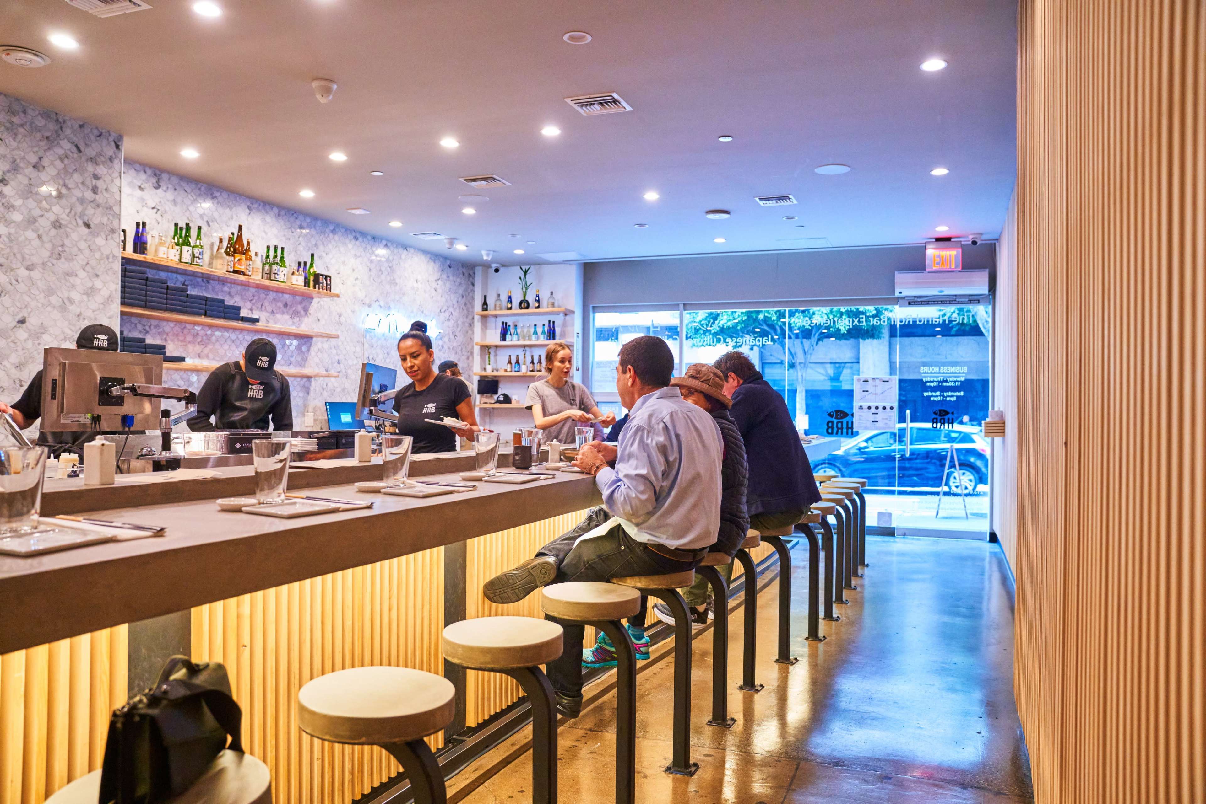 A modern restaurant interior with a bar counter where several patrons are sitting, and staff preparing drinks in the background.