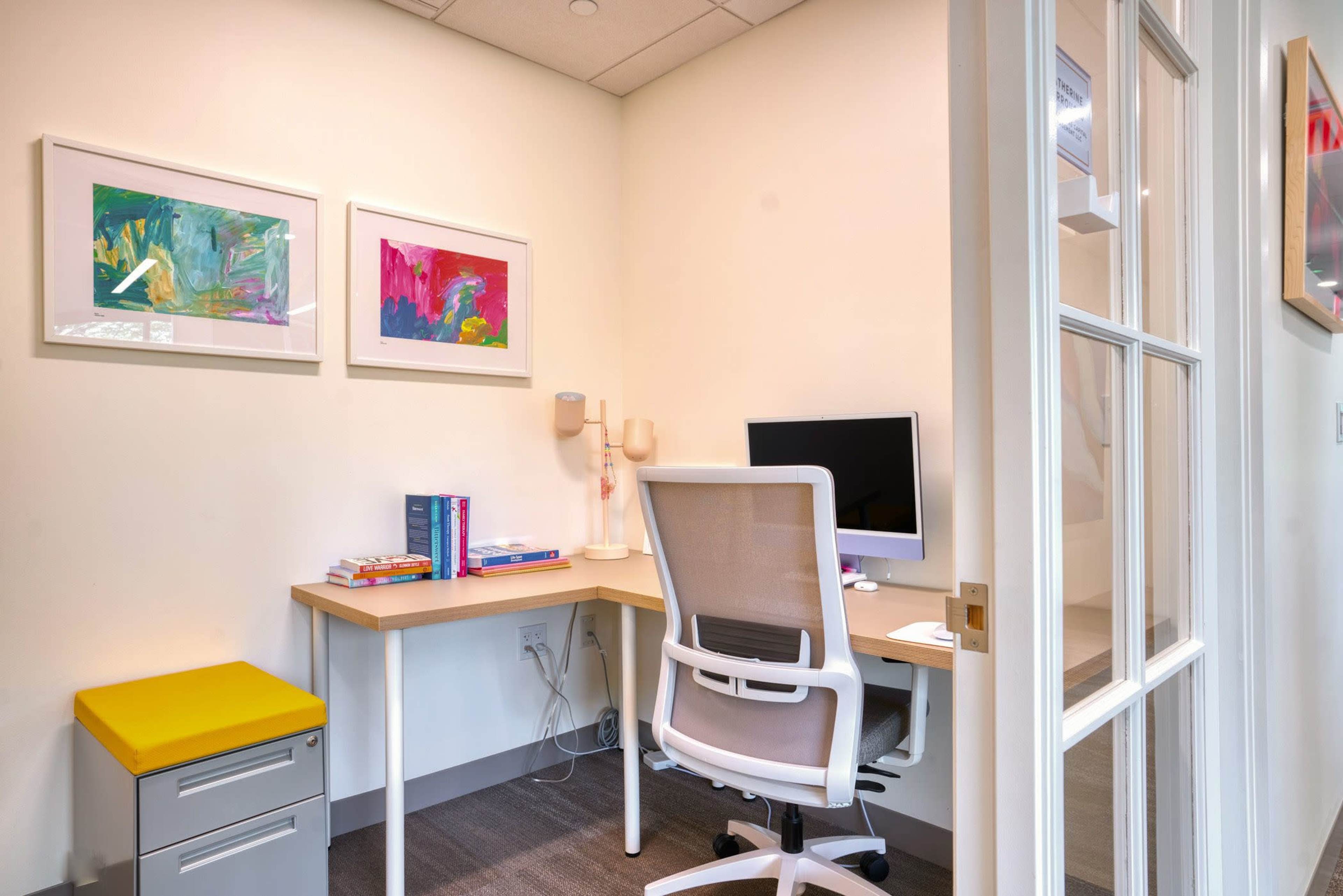 A bright office space features a desk with a computer, books, and artwork on the walls, complemented by a modern chair and a yellow storage unit.