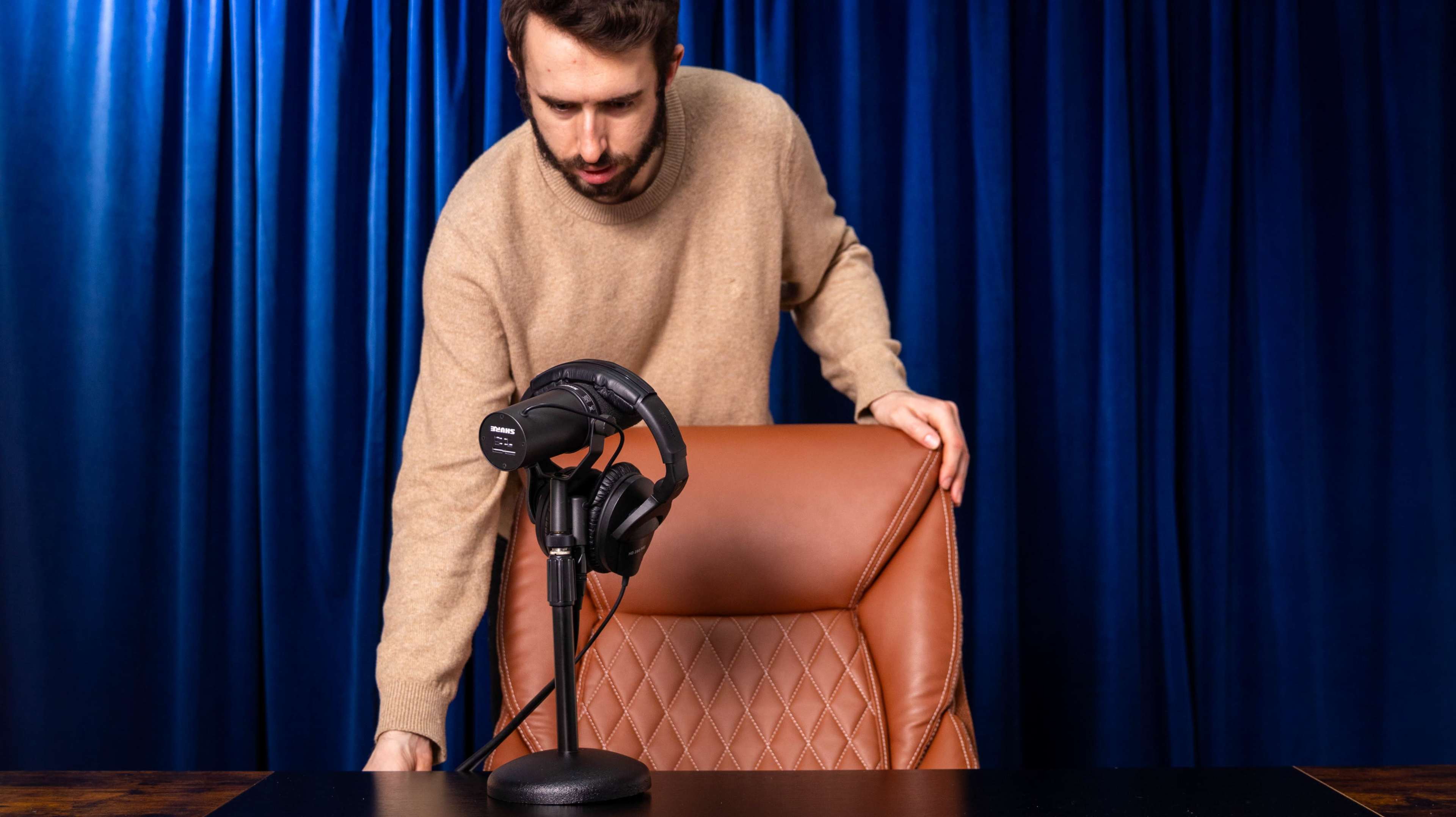 A man in a beige sweater is adjusting a microphone on a desk with a brown chair against a blue backdrop.