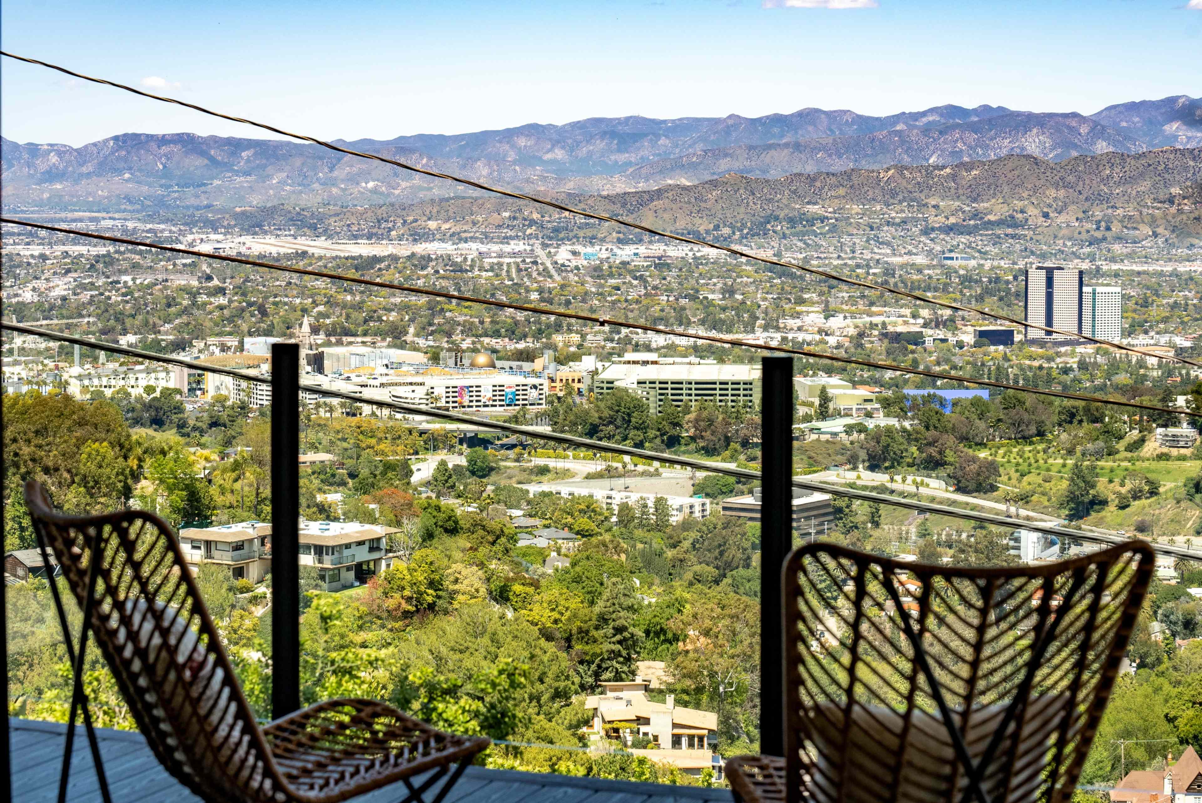 A panoramic view of a lush green landscape and urban development from a balcony with two wicker chairs.
