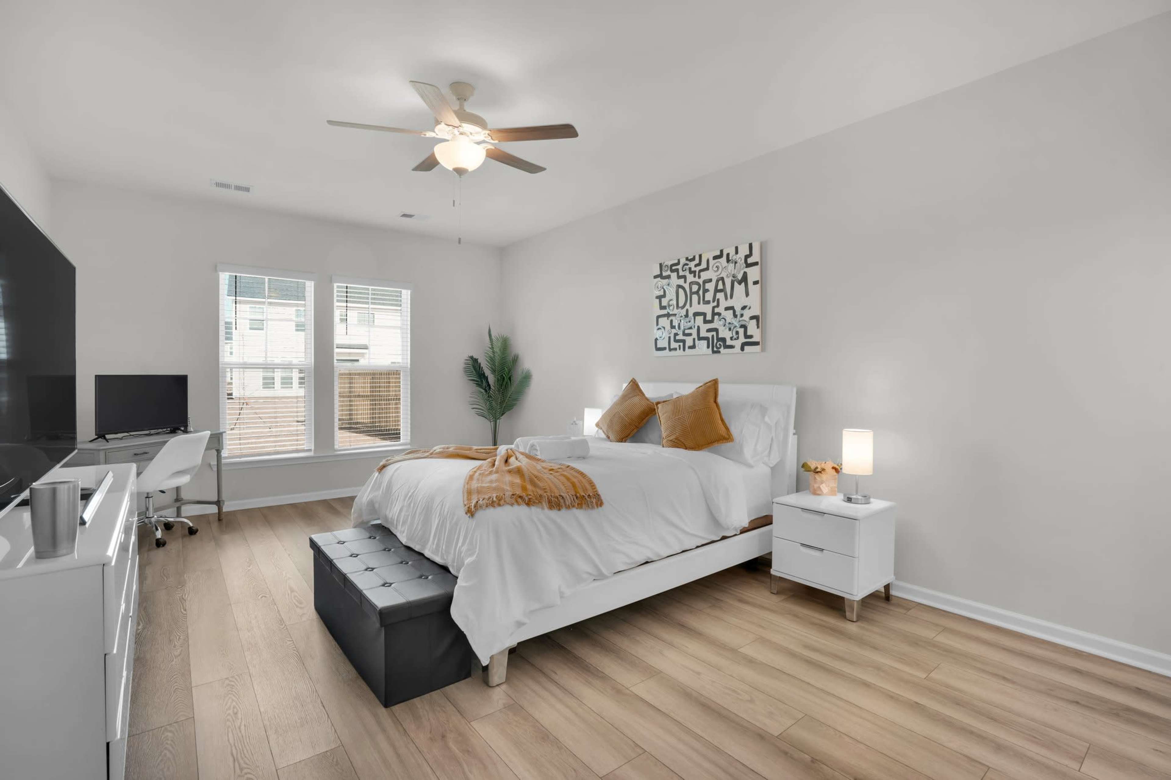 The image shows a well-lit bedroom featuring a bed with white linens and decorative pillows, a bedside table with a lamp, a bench at the foot of the bed, and a television on a stand across from the bed.