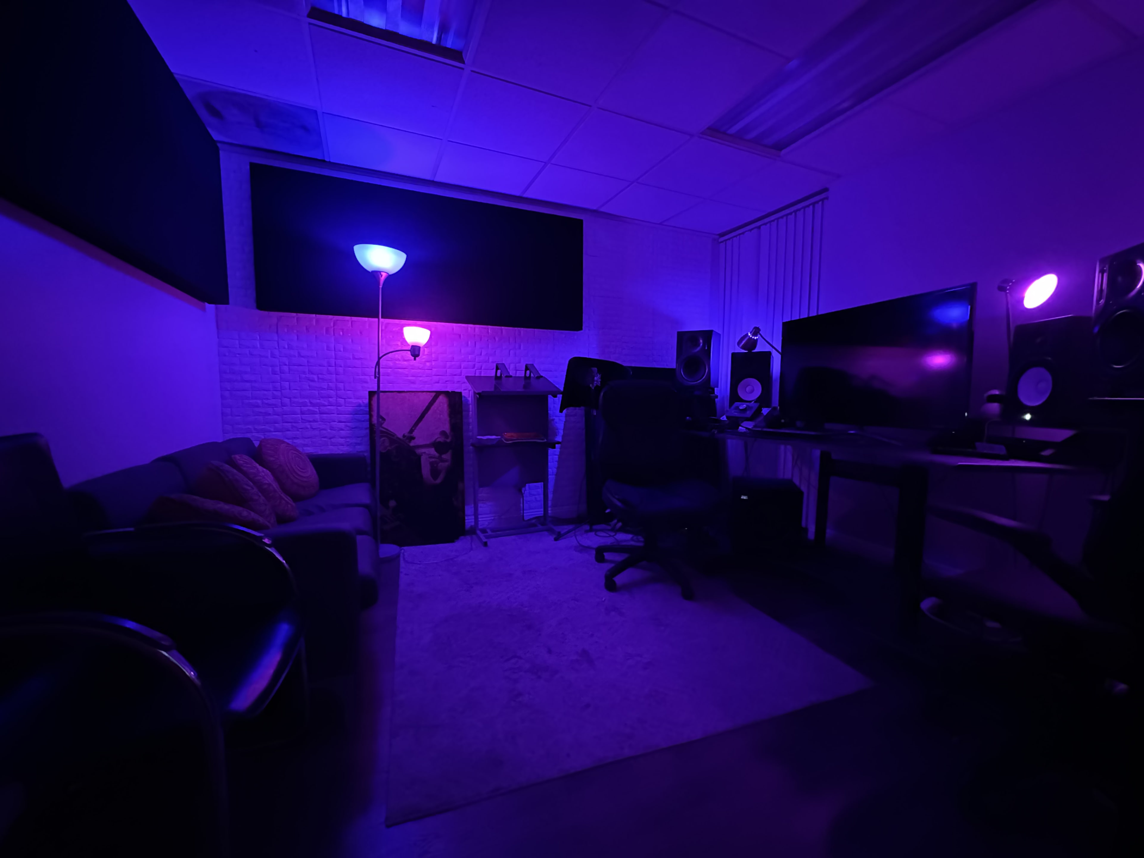 A dimly lit room features a couch, a desk with monitors, and colorful lighting from lamps, creating a workspace atmosphere.