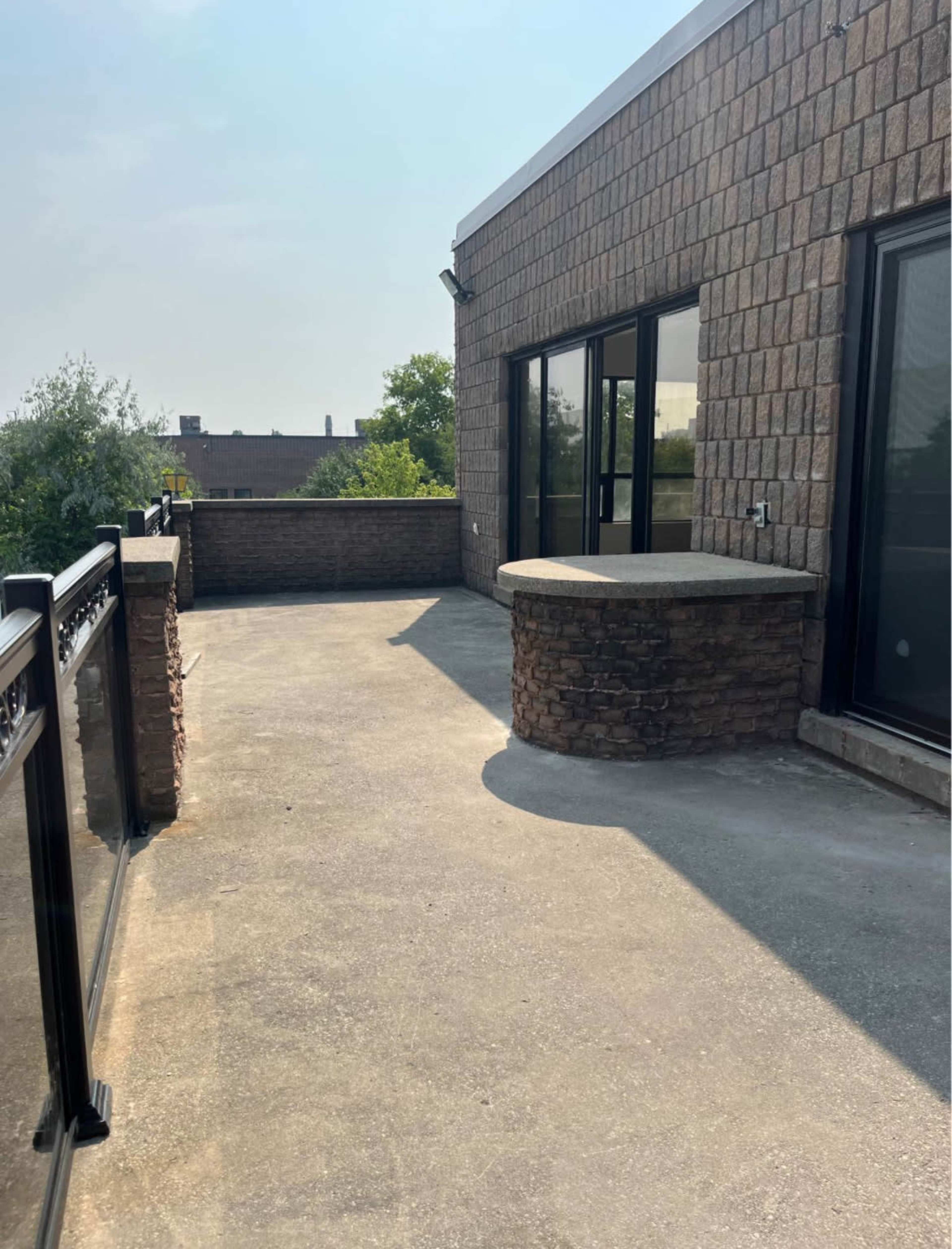A spacious patio with a stone bar counter and glass railing, surrounded by green trees and a clear sky.