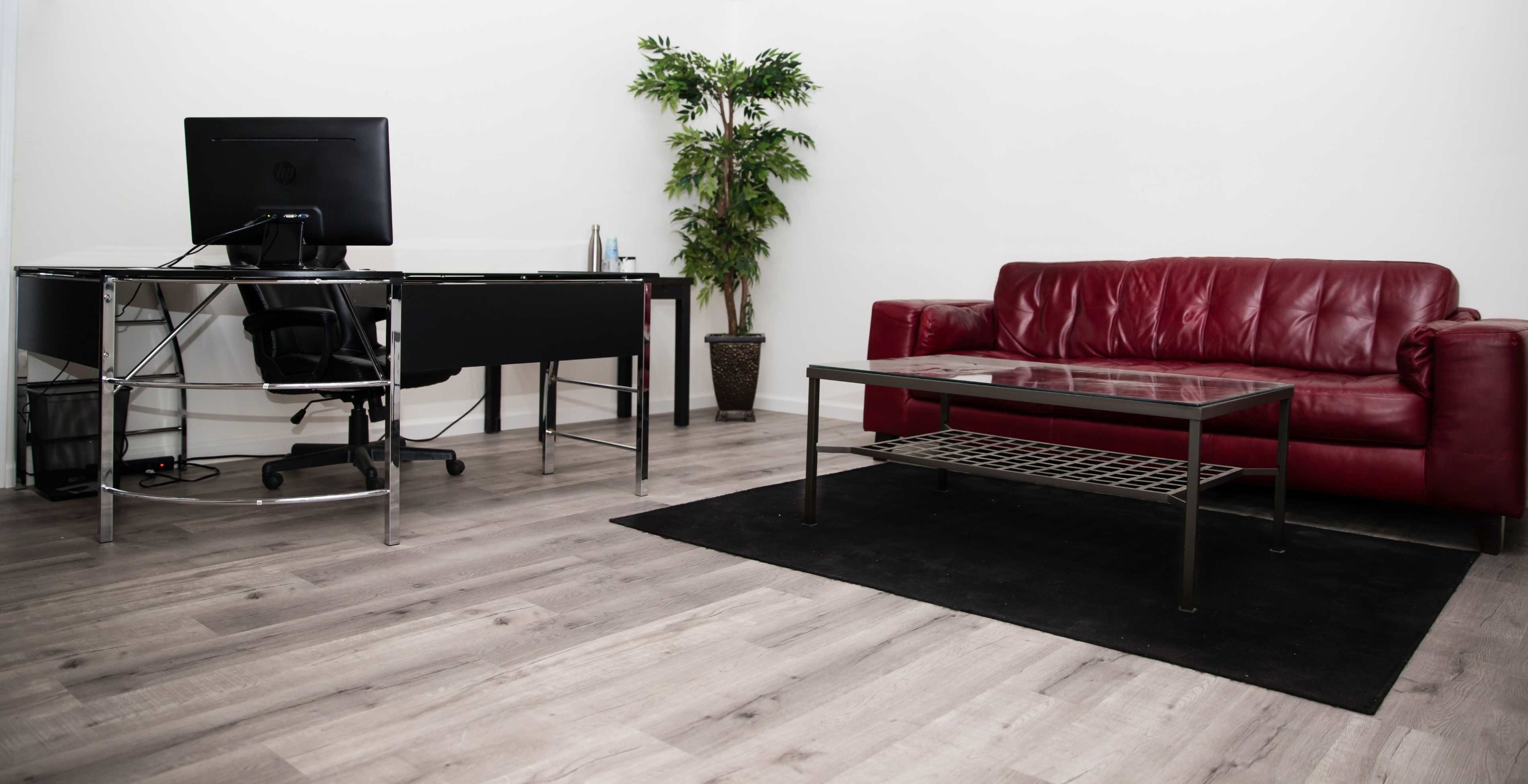 A modern office space featuring a desk with a computer, a red leather sofa, a glass coffee table, and a decorative plant in a pot.