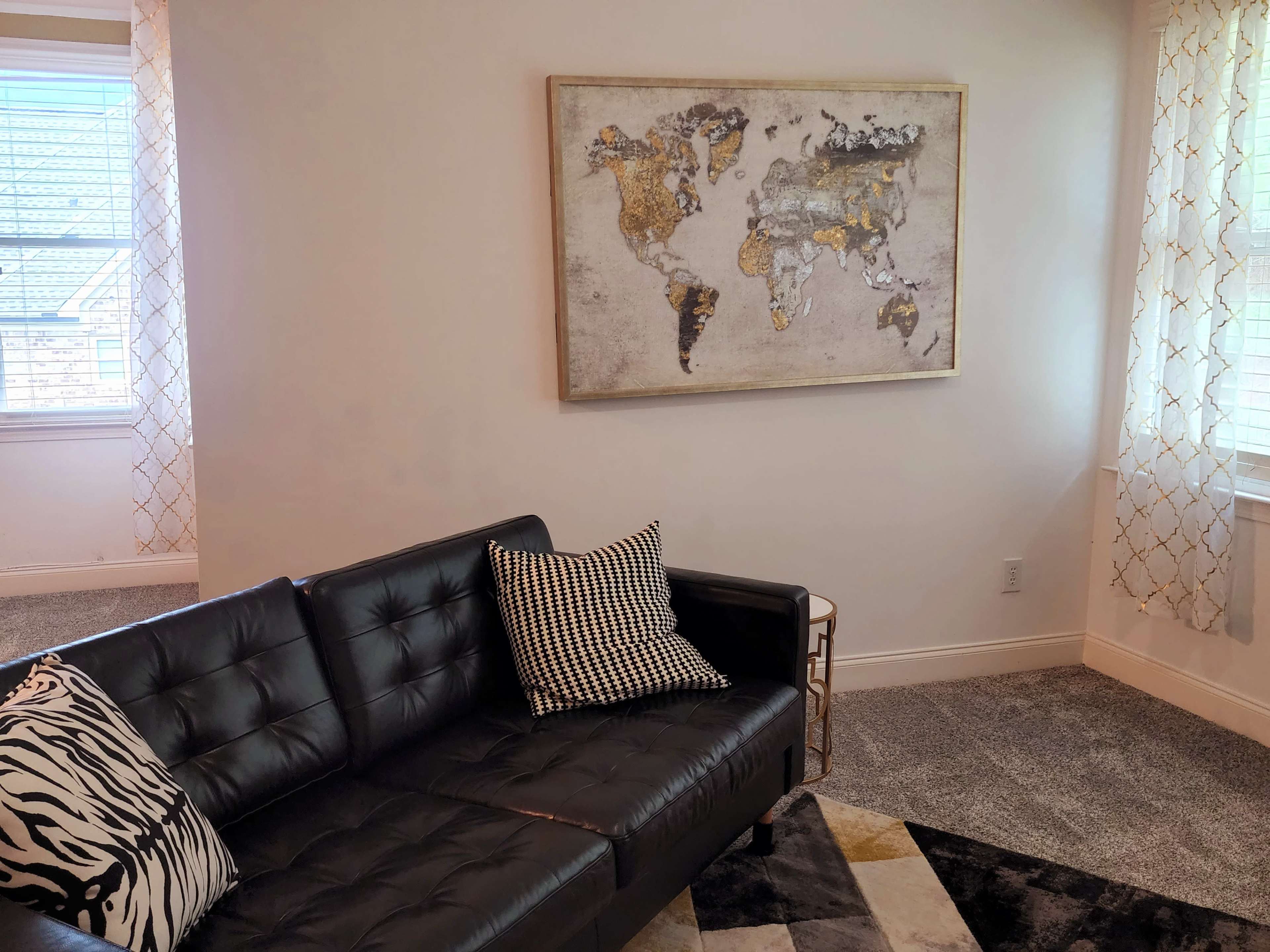 A black leather sofa with a patterned pillow faces a wall featuring a gold-accented world map, under soft lighting from nearby windows.