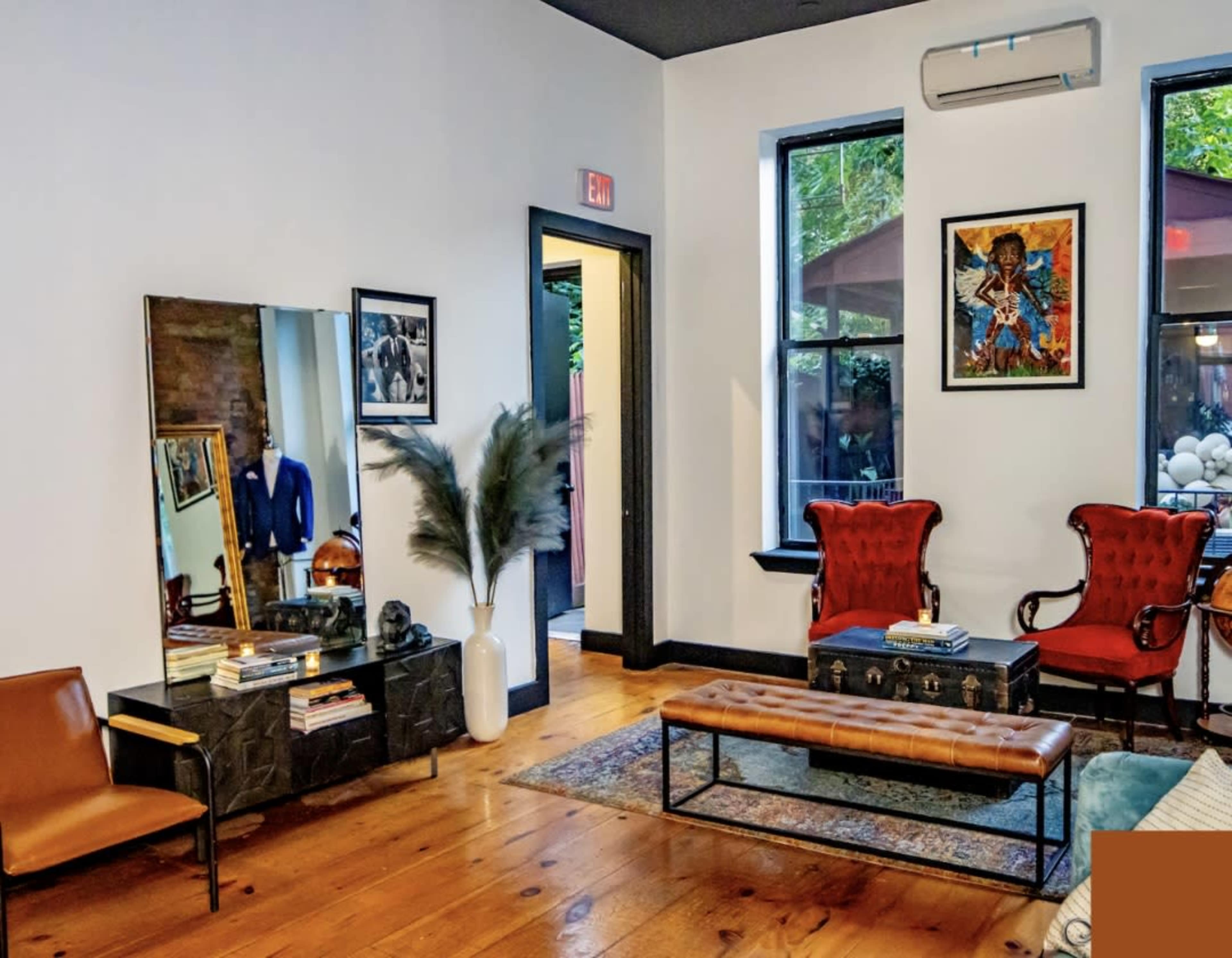 The image shows a modern living room with wooden floors, two red armchairs, a brown leather bench, a large mirror, and artwork on the walls.