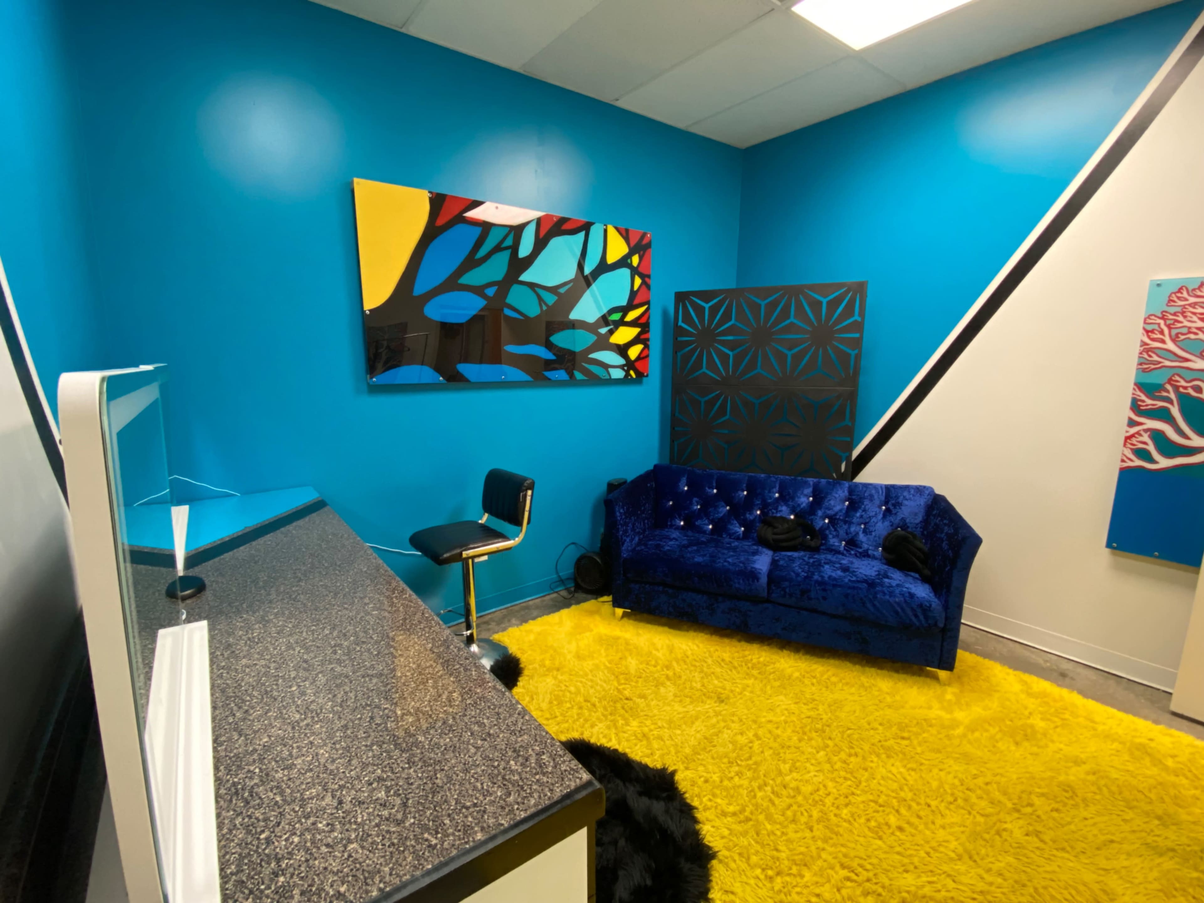 The room features a bright blue wall, a blue velvet couch, a black chair, and a yellow rug, with decorative artwork displayed on the walls.