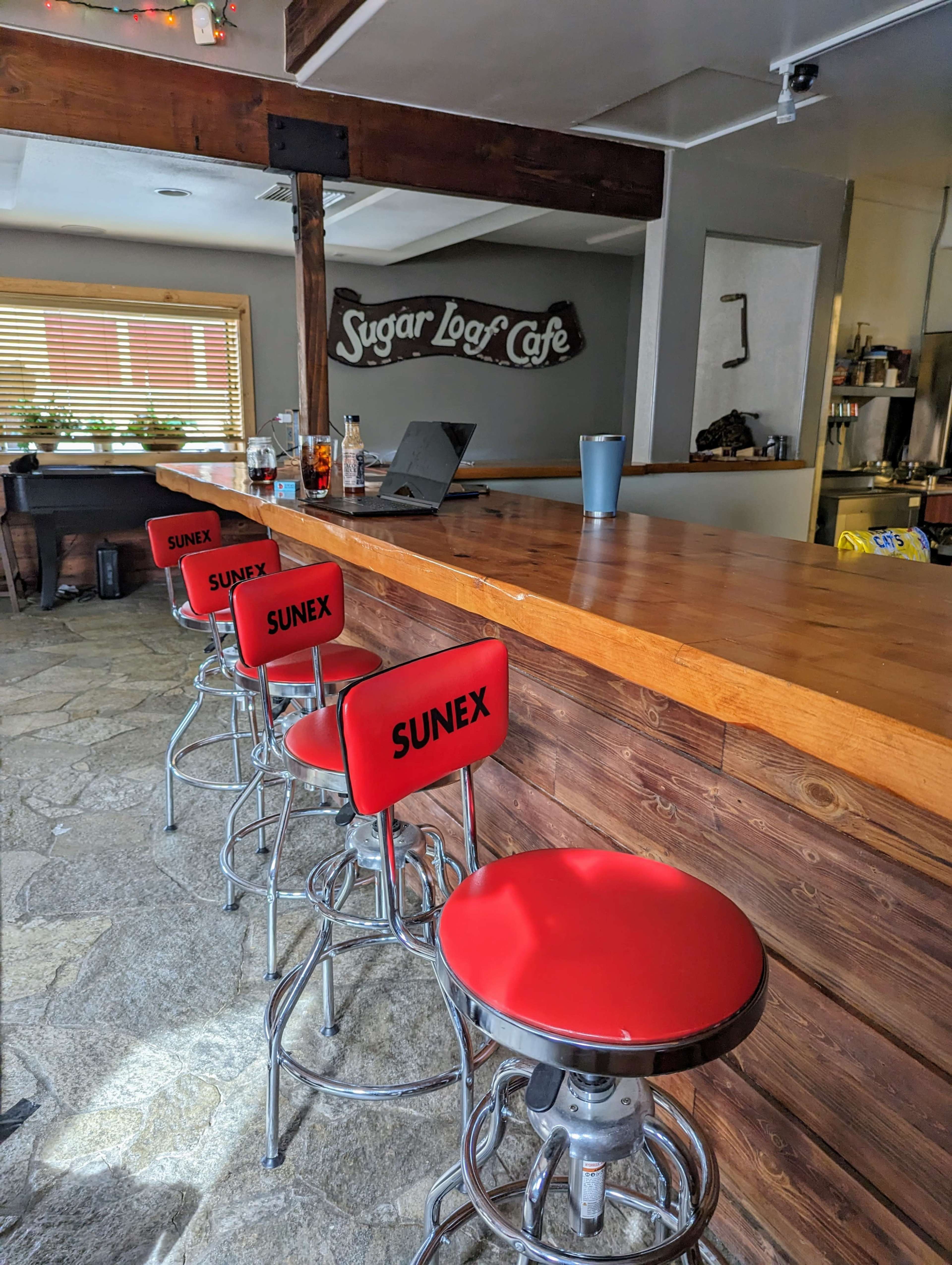 A wooden bar with four red stools in a café setting, featuring a sign that reads "Sugar Logs Café" in the background.