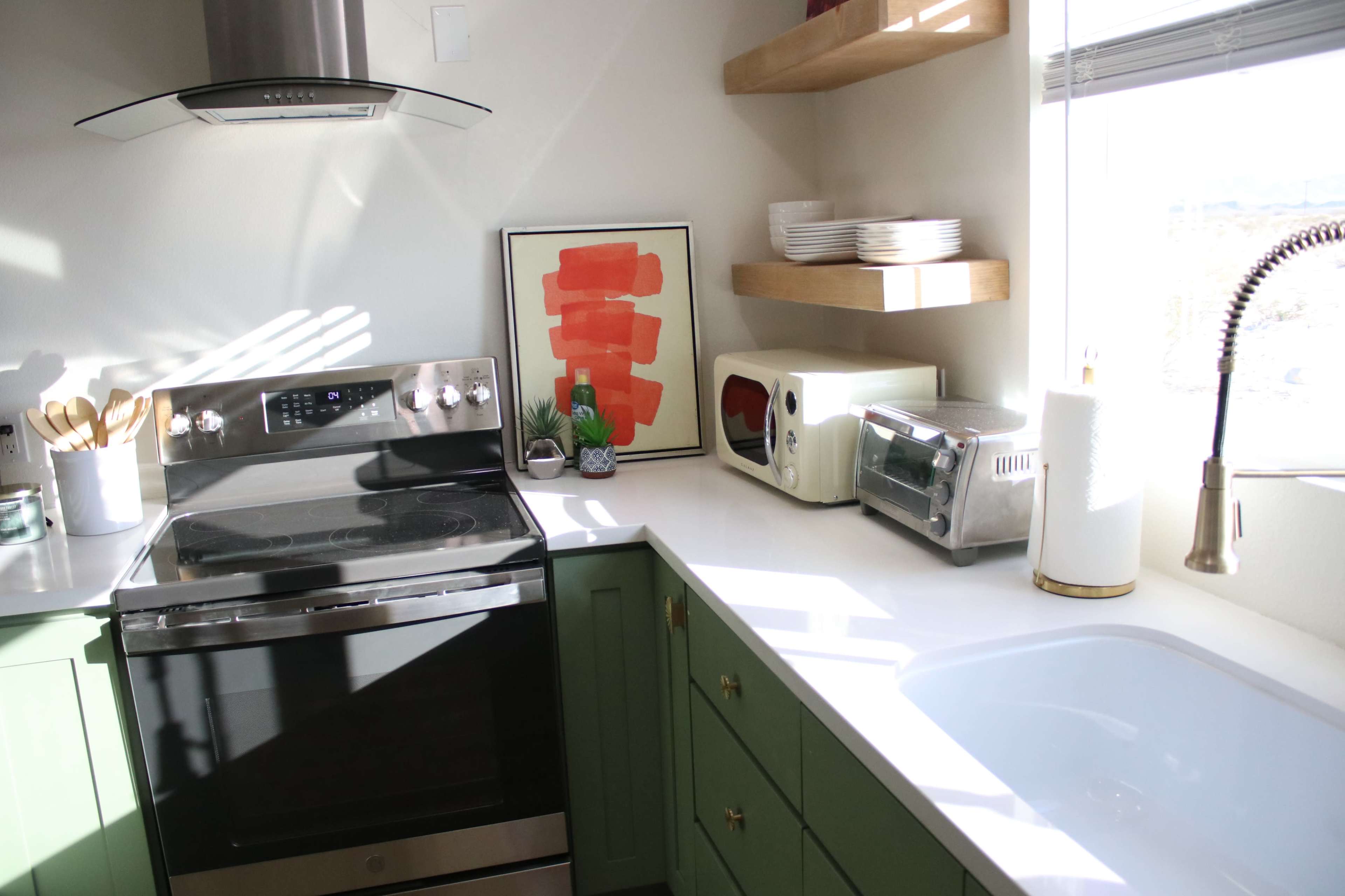 A modern kitchen features a stove, sink, and various appliances, with open shelves displaying plates and a framed artwork.