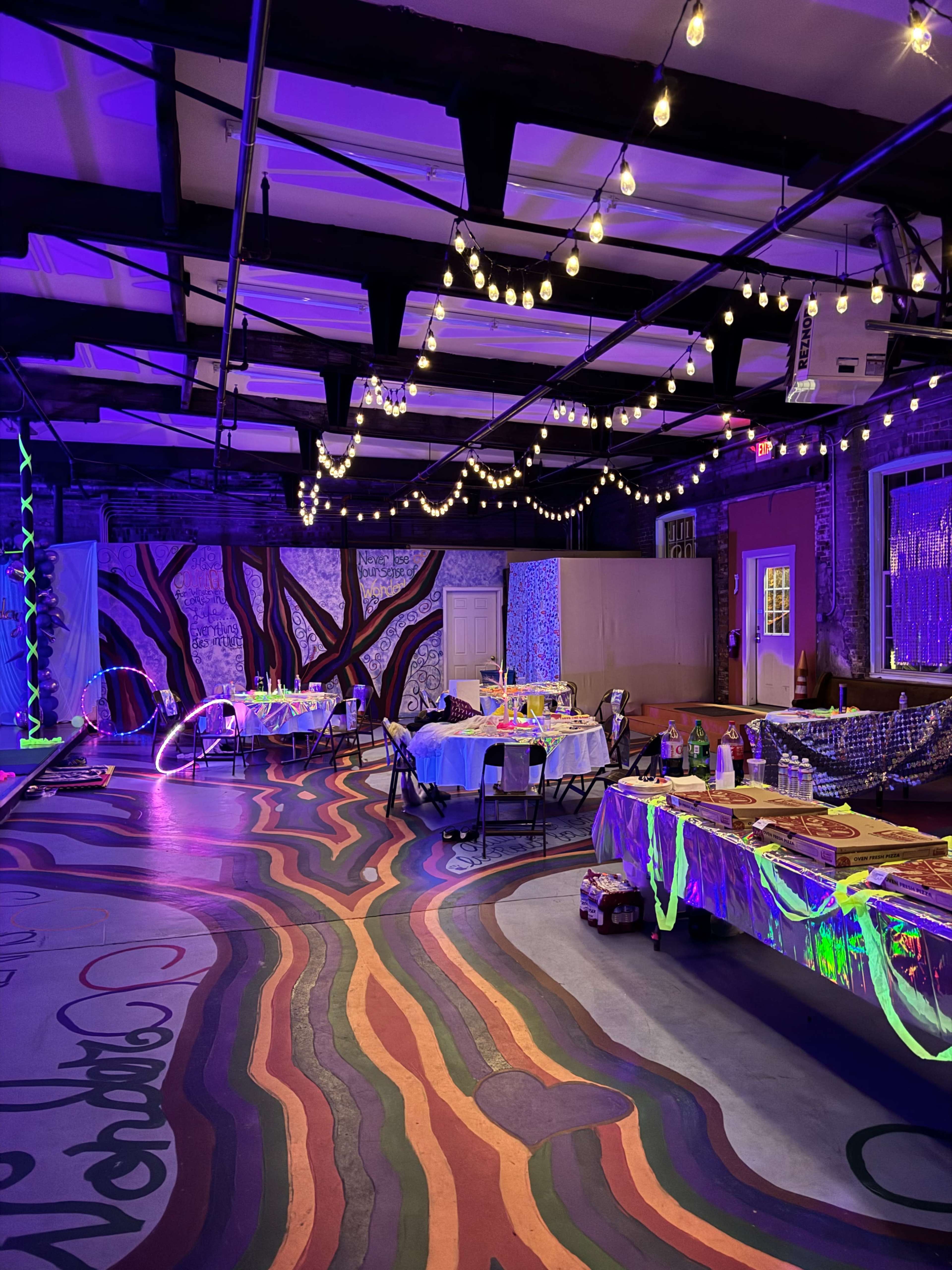 A dimly lit event space decorated with colorful tables under string lights and featuring vibrant, wavy floor patterns.