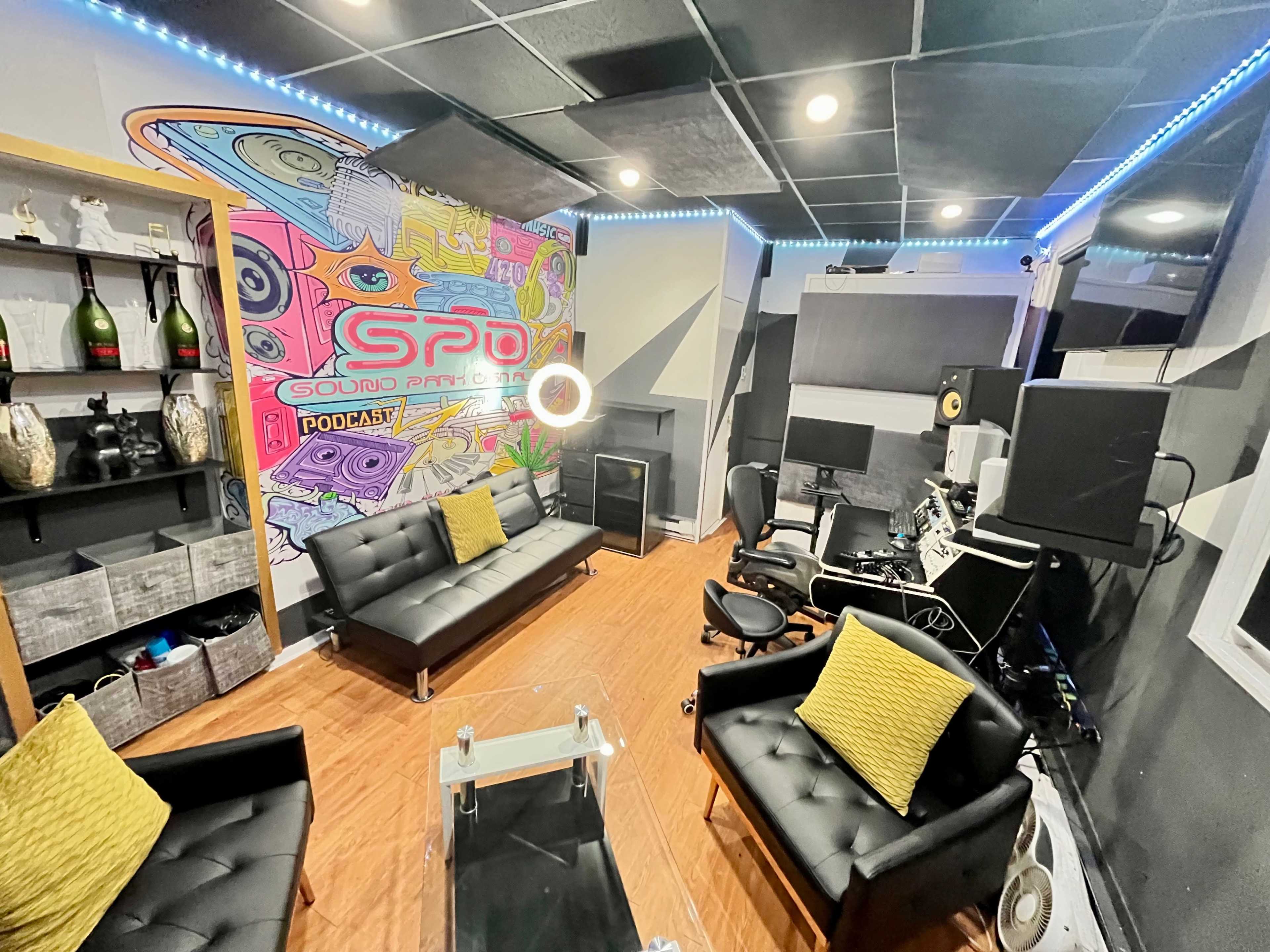 The image shows a podcast recording studio featuring a colorful mural, black furniture, and audio equipment.