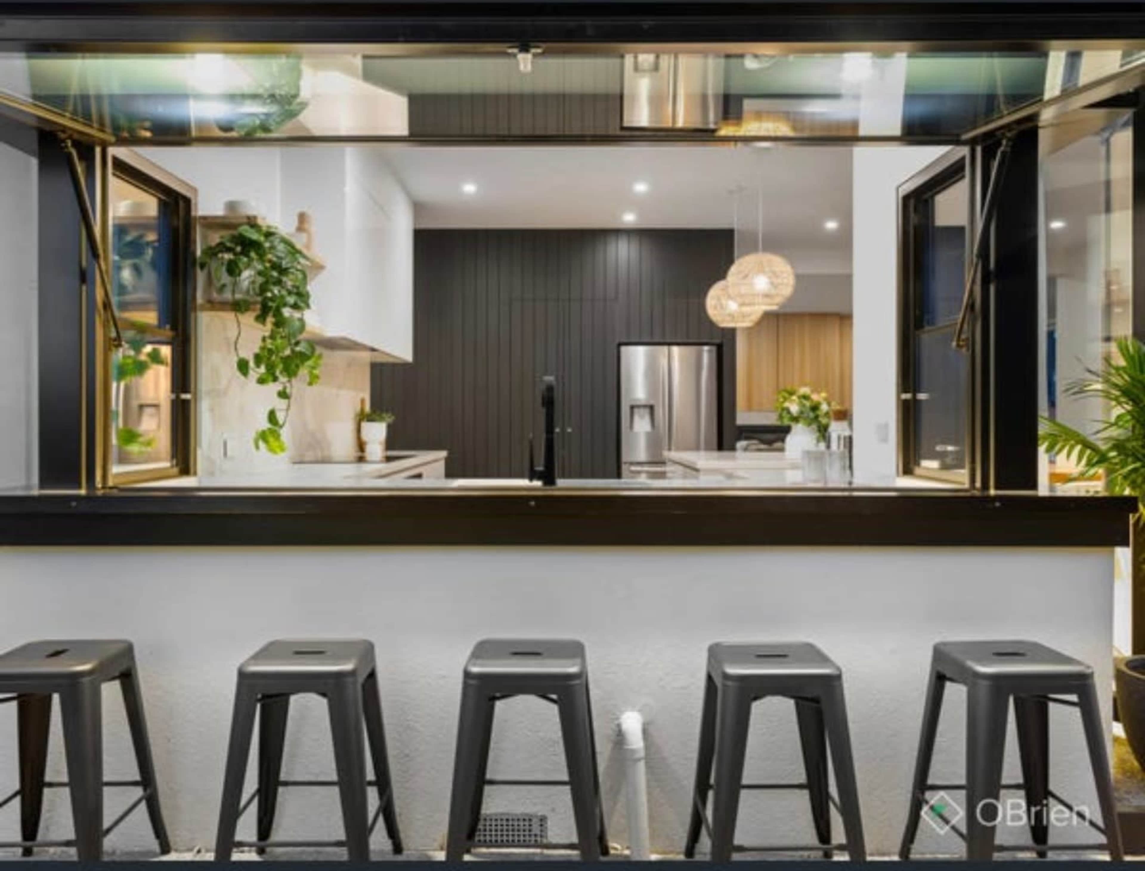 The image shows a kitchen with a large open window overlooking a row of black metal stools placed outside.