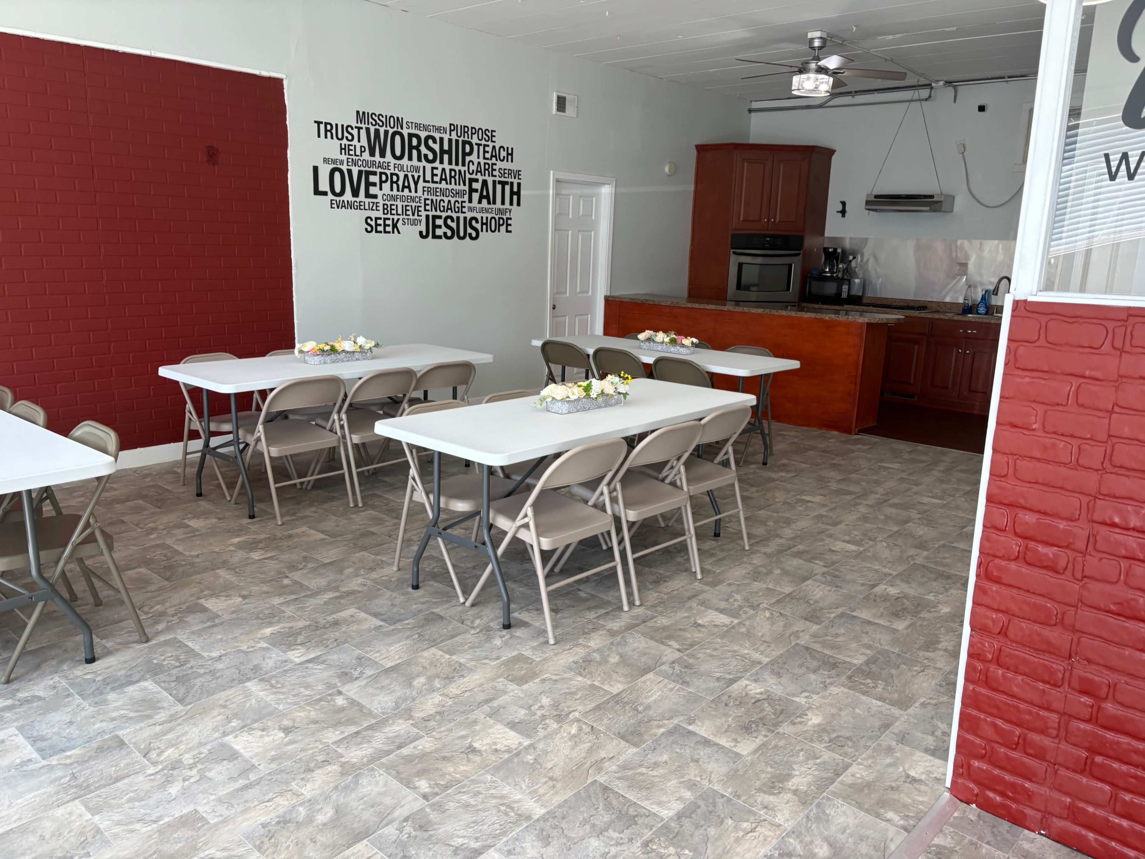 The image shows a room with tables arranged for an event, featuring a kitchen area with wooden cabinetry and motivational wall decals.