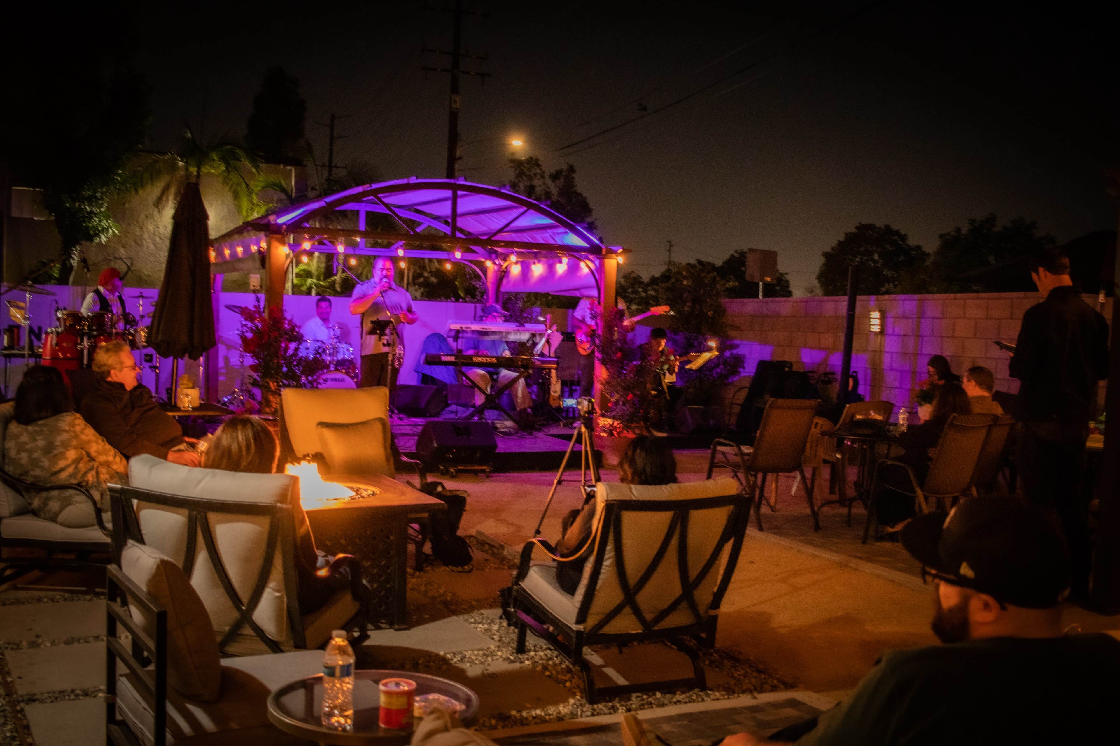 A live band performs under a lit gazebo in a backyard while guests relax around fire pits.