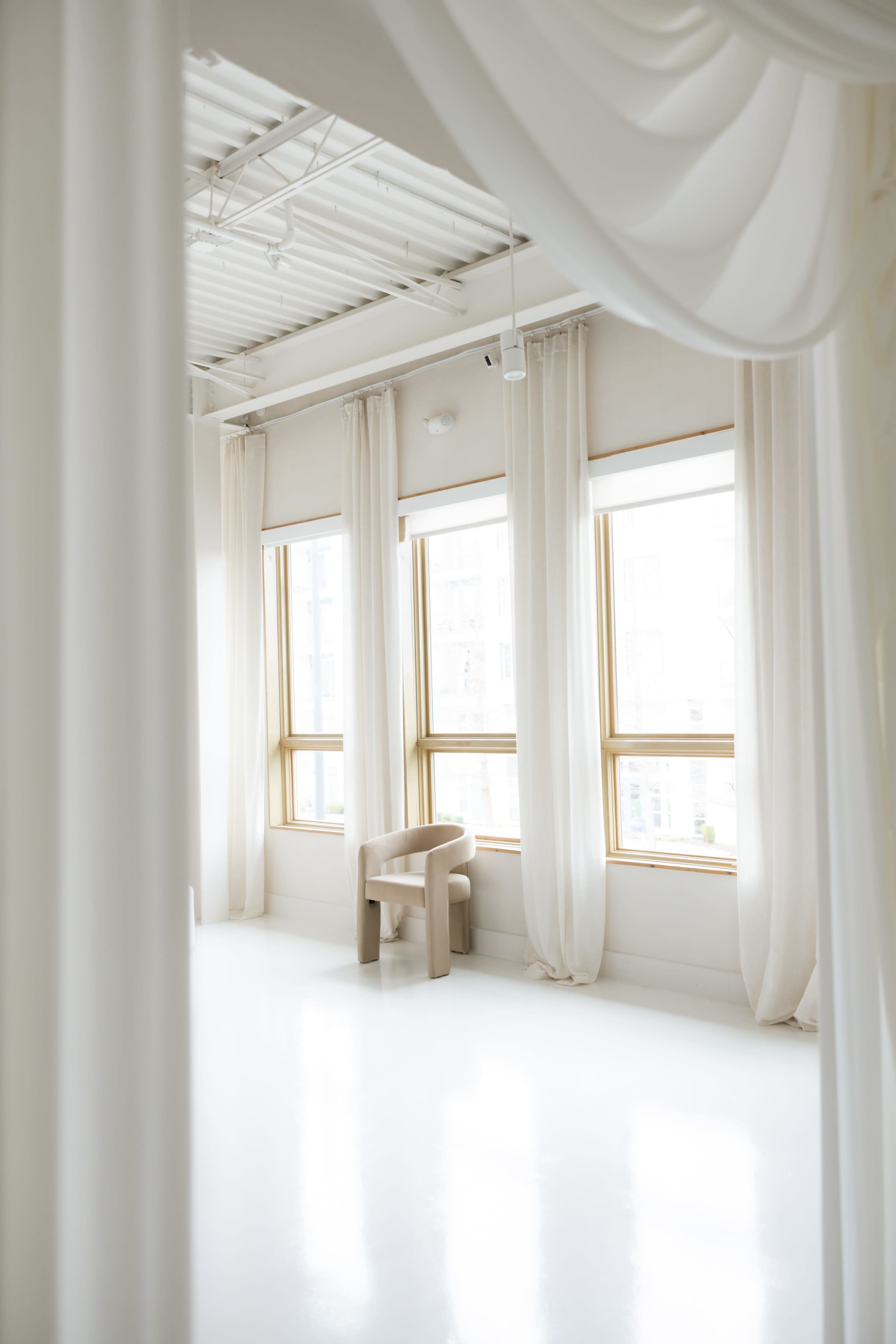 The image shows a bright, minimalistic room with large windows adorned with sheer curtains and a simple wooden chair.