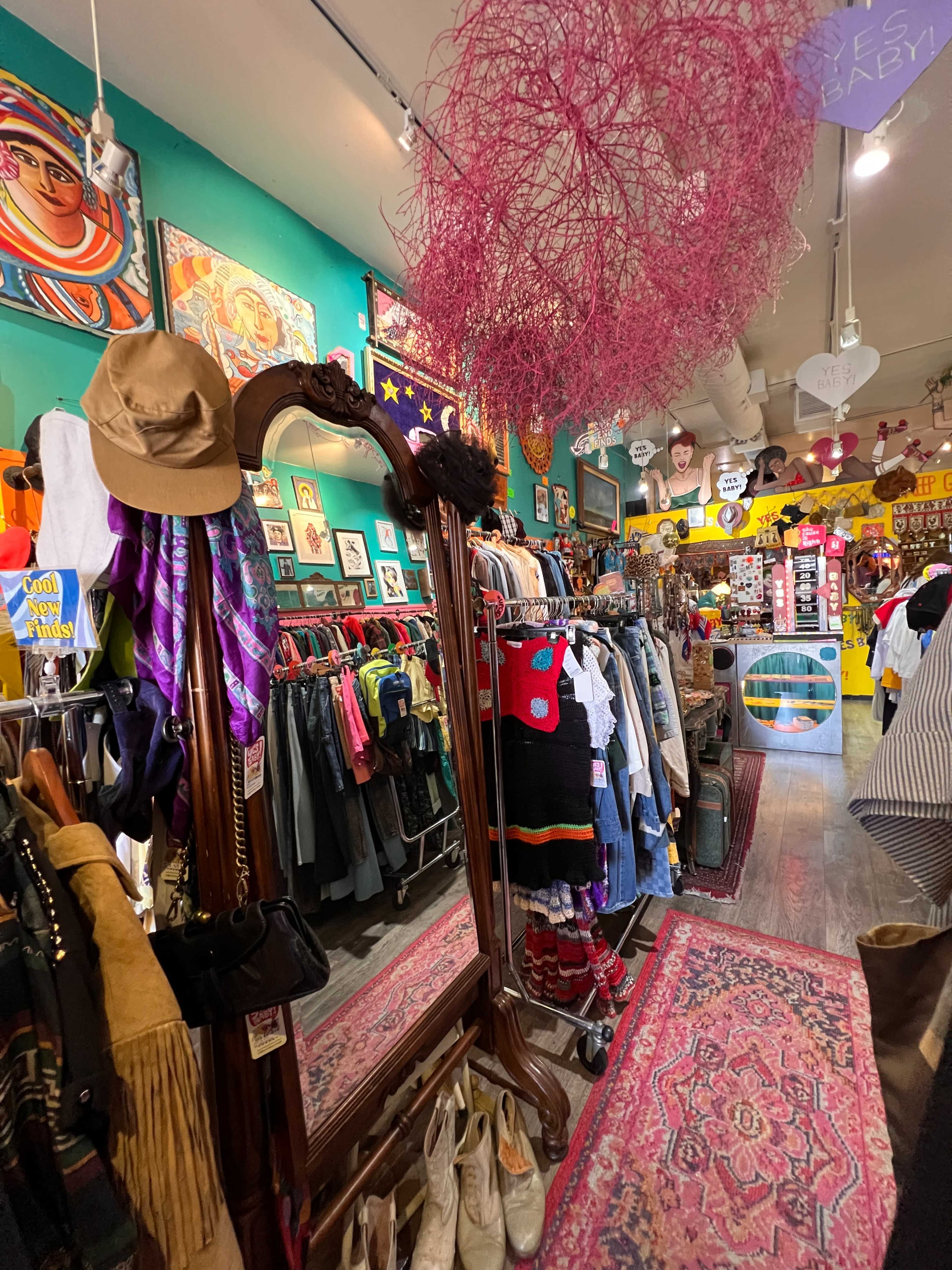The image shows a colorful vintage clothing shop filled with racks of clothes, accessories, and artwork, featuring a large mirror reflecting the vibrant interior.
