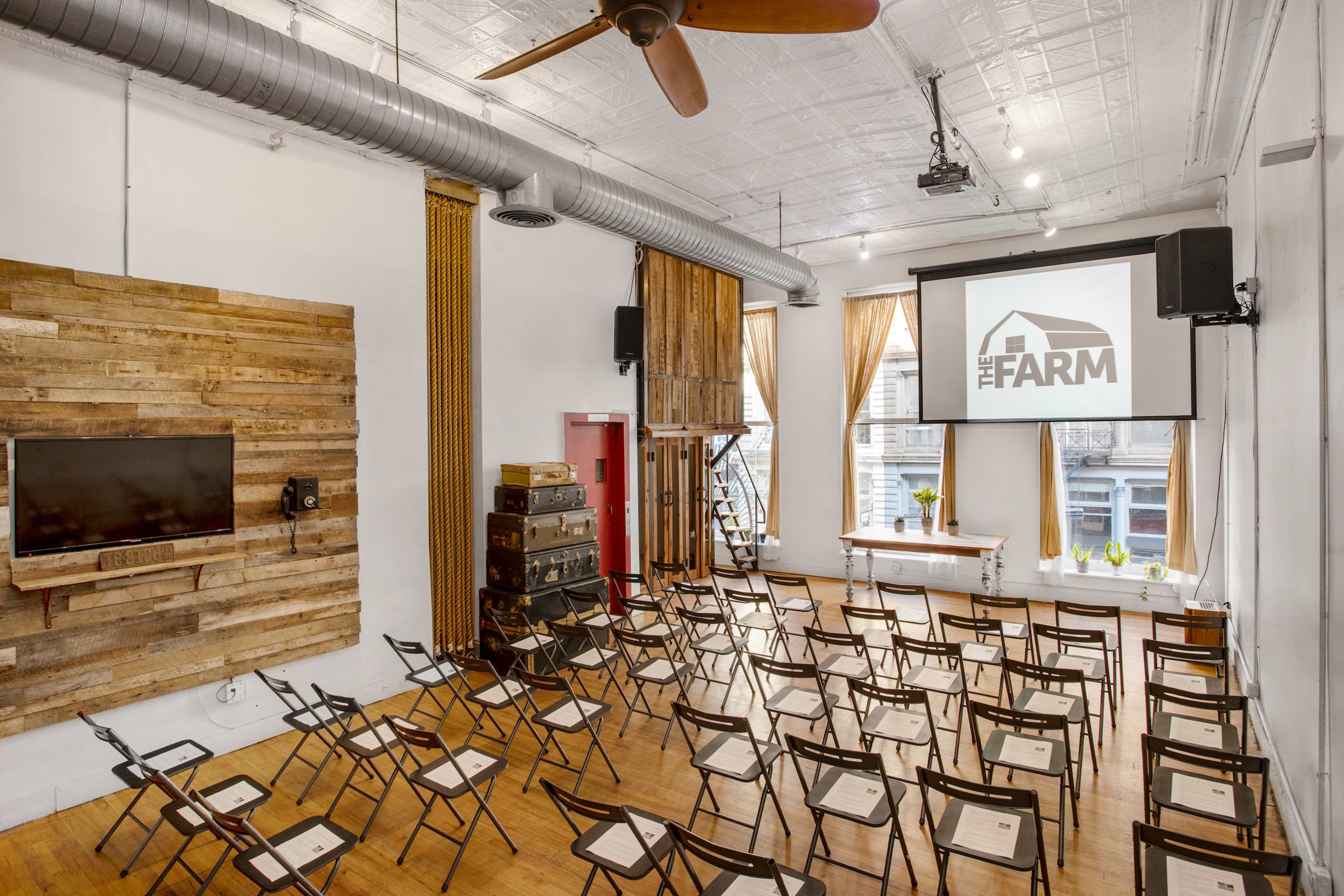 A seminar room with rows of folding chairs arranged in front of a projector screen, wooden paneling on one wall, and large windows providing natural light.
