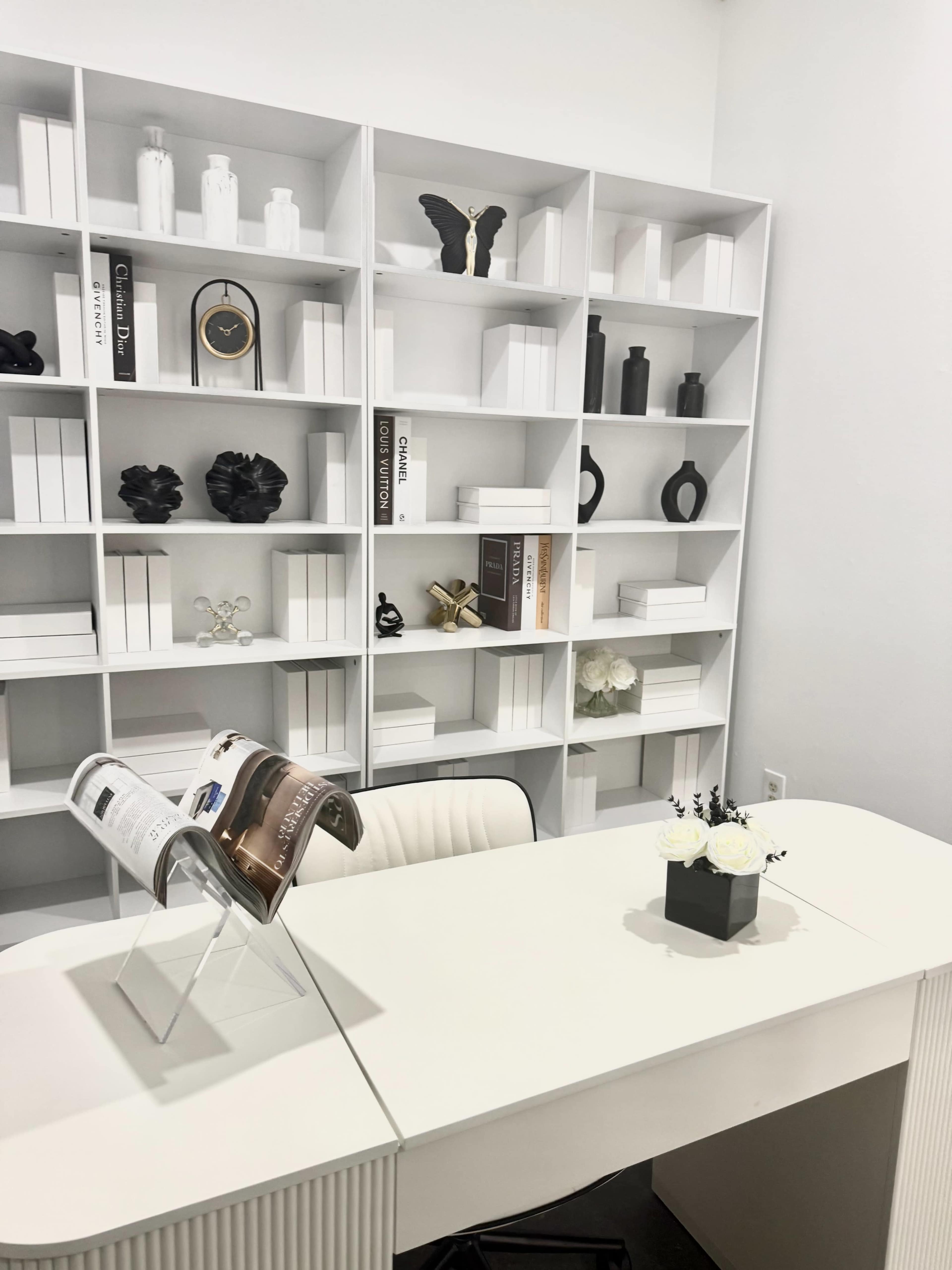A minimalist office space features a white desk with an open magazine stand and a decorative floral arrangement, set against a white shelf displaying a variety of objects and books.