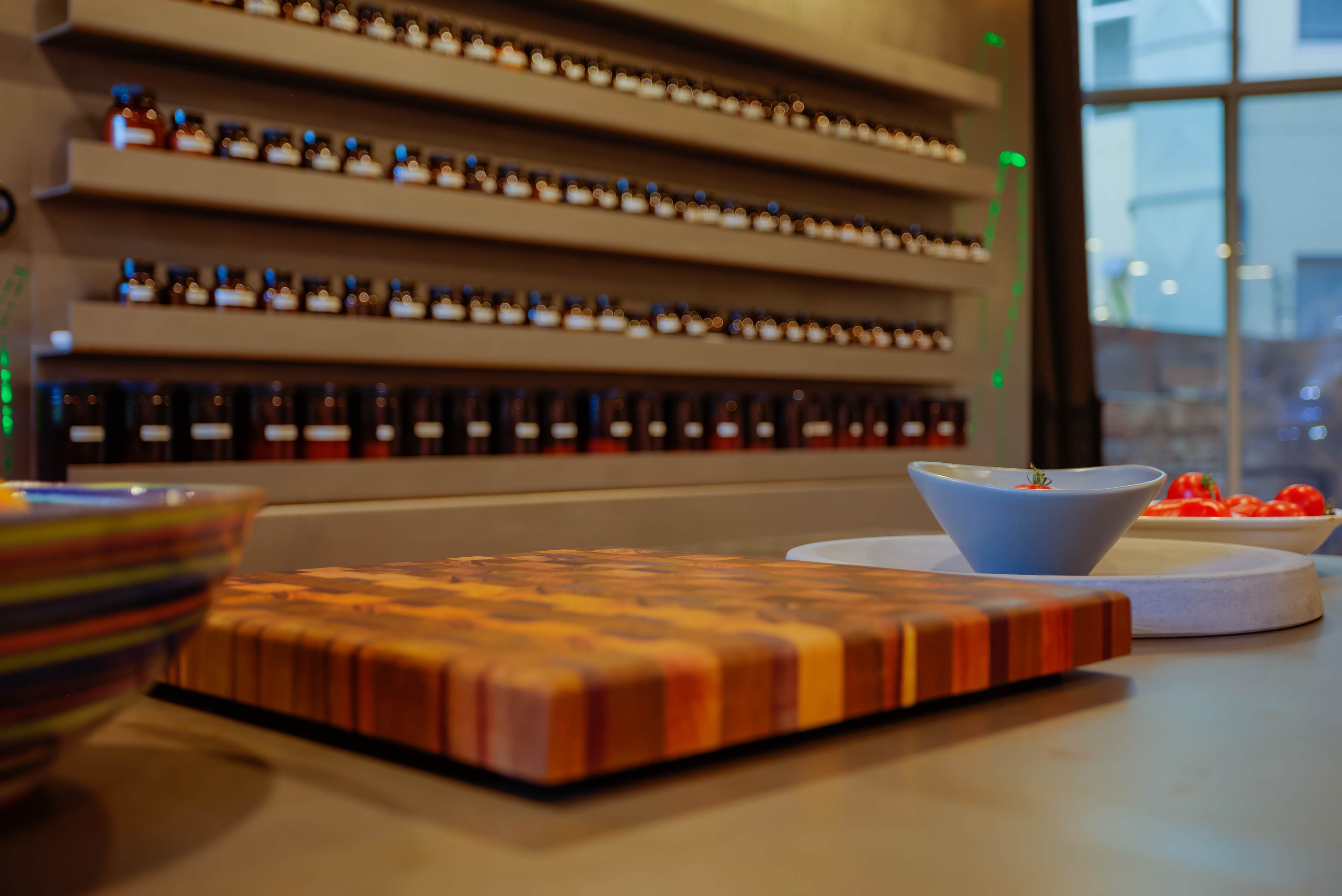 A wooden cutting board placed on a countertop with bowls and fresh tomatoes in the foreground, against a backdrop of shelves displaying numerous jars.
