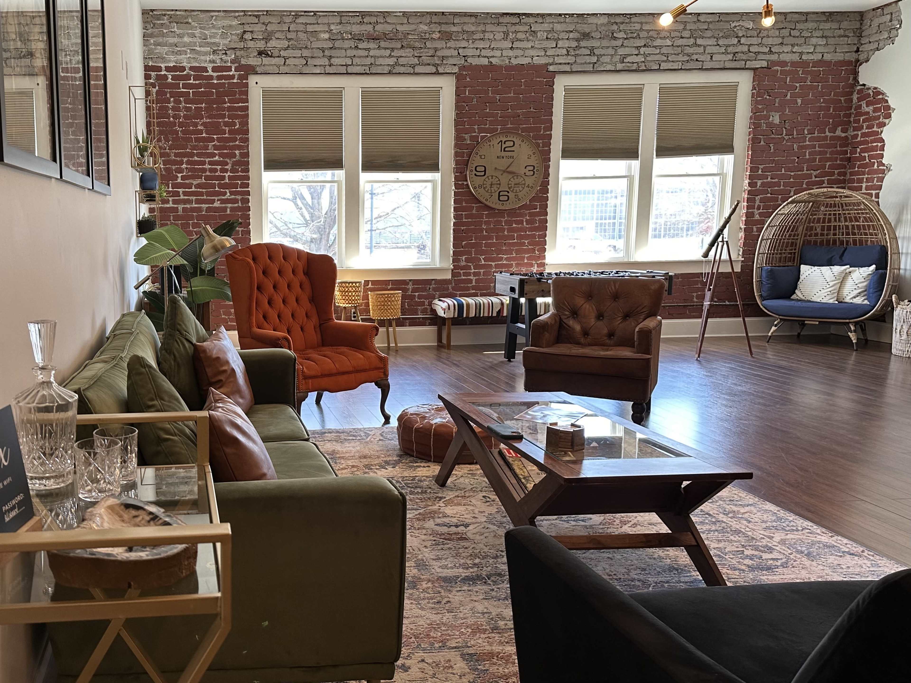 A spacious room with exposed brick walls, various seating options including orange and brown armchairs, a glass coffee table, and large windows letting in natural light.