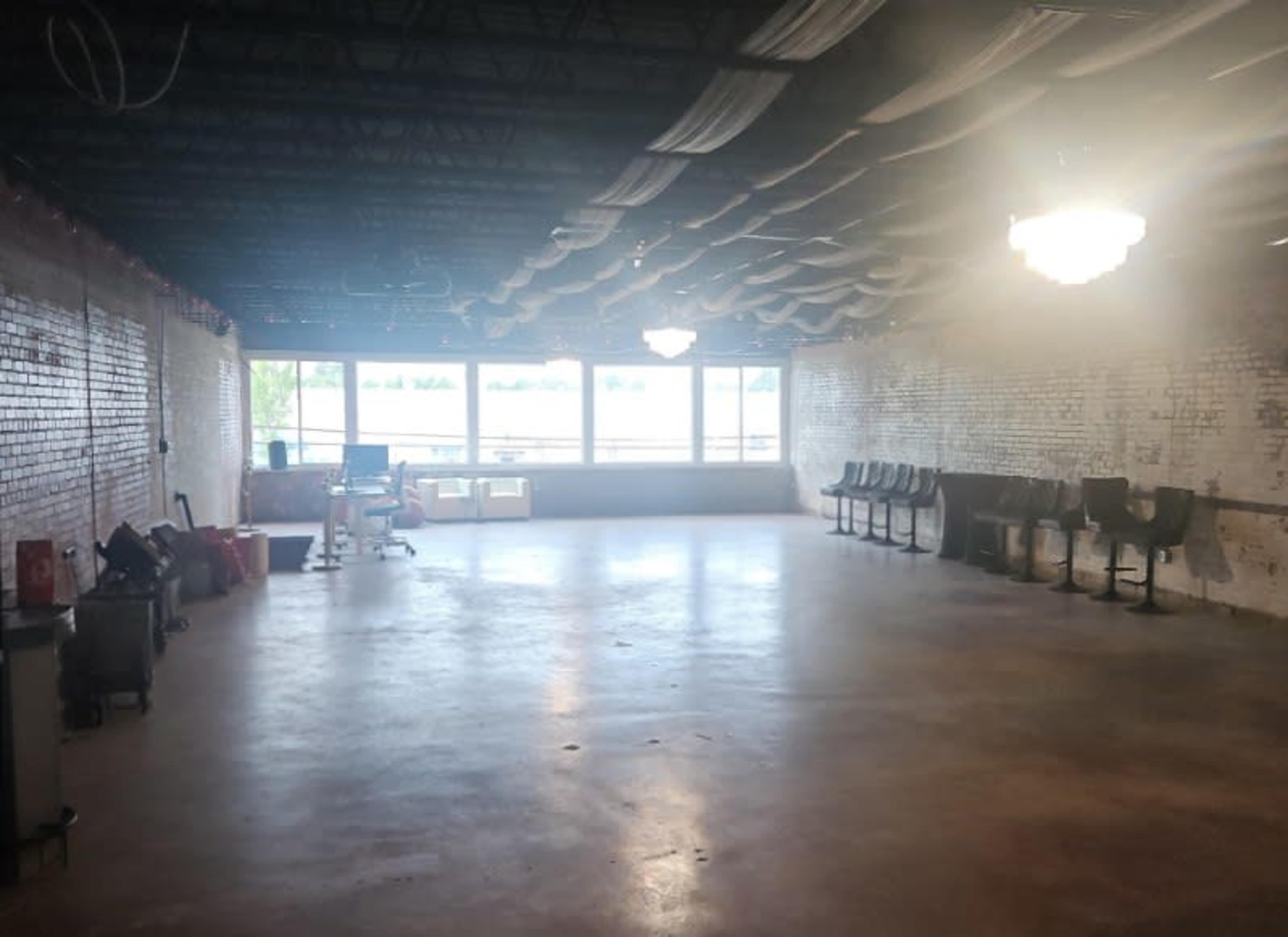 The image shows a spacious, empty room with exposed brick walls, concrete flooring, and large windows allowing natural light to enter.