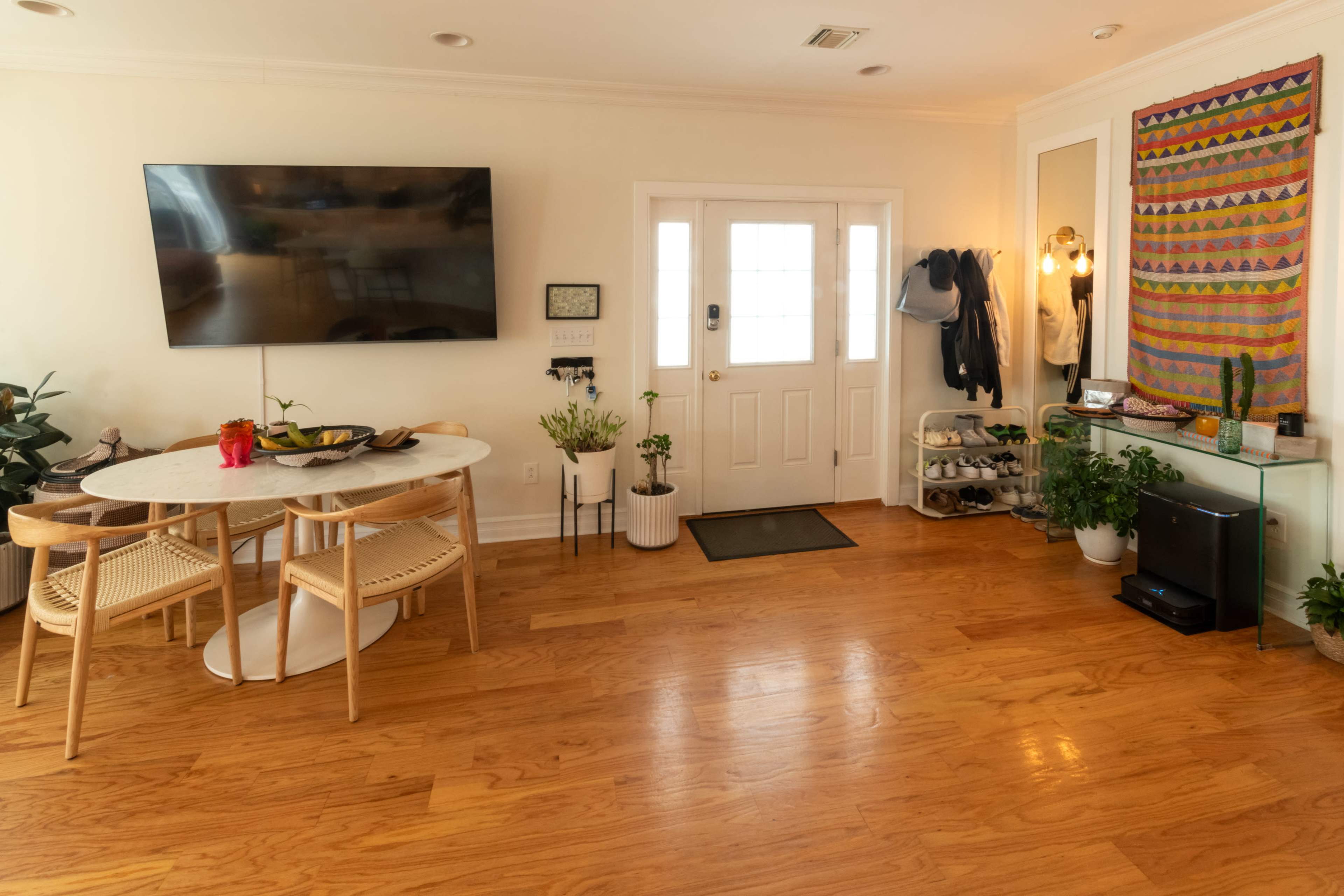 The image shows a bright and modern living space with a round dining table, a television mounted on the wall, and an entrance door flanked by plants and decorative items.