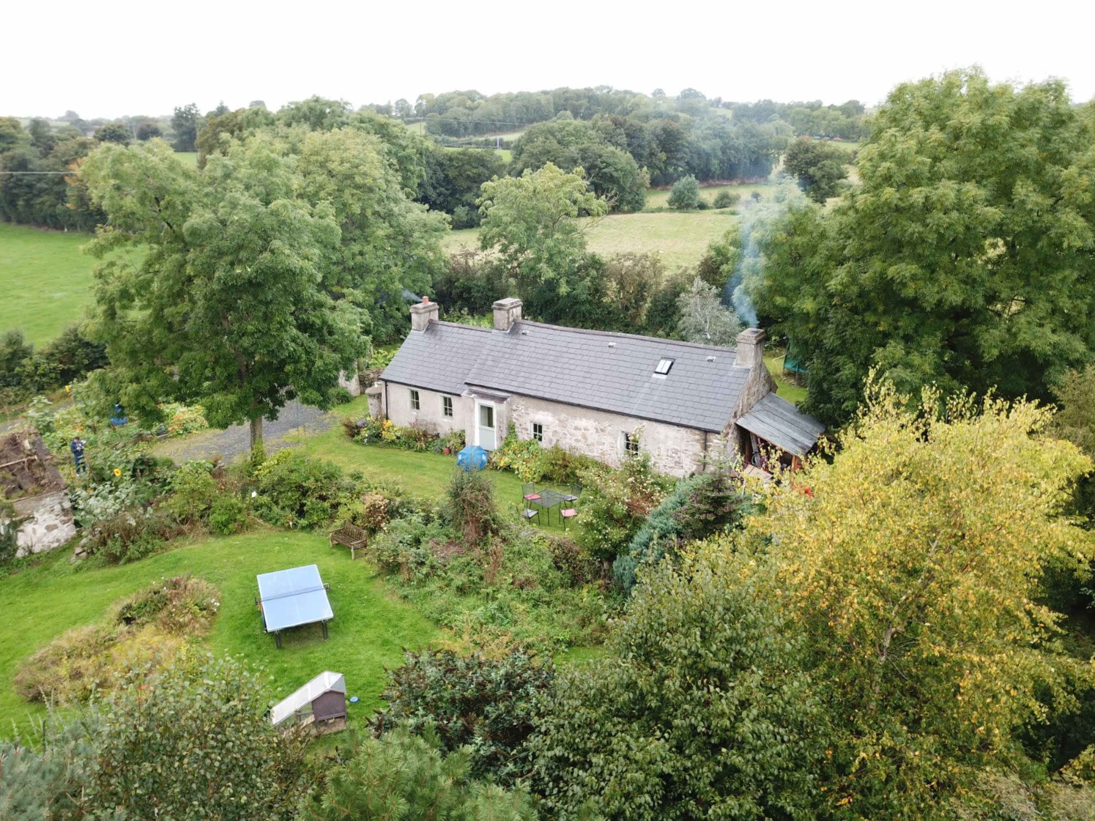 An aerial view shows a stone cottage surrounded by lush greenery and trees, with smoke rising from its chimney.