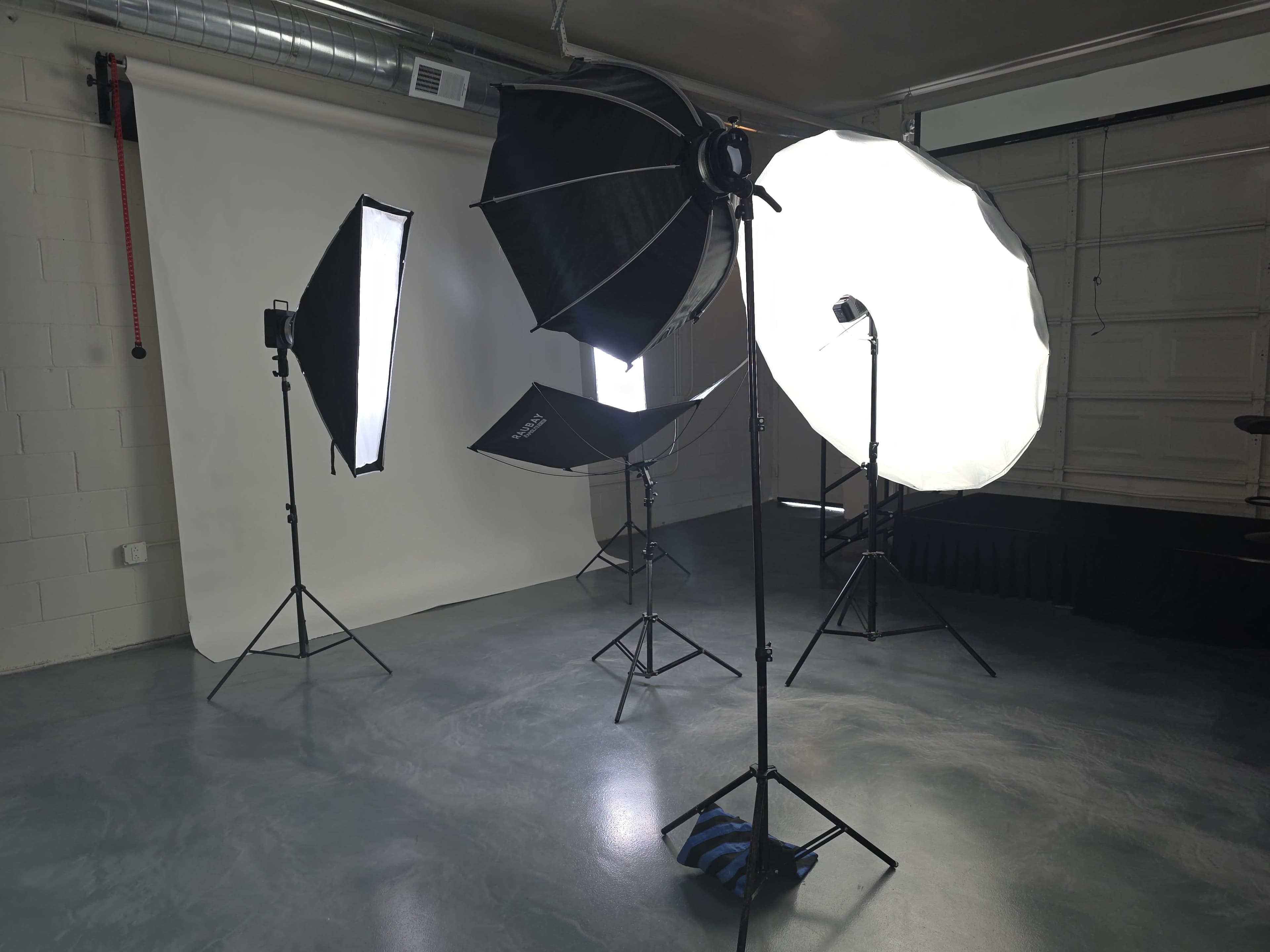 The image shows a professional photography studio with multiple lighting setups, including softboxes and reflectors, positioned around a seamless backdrop.