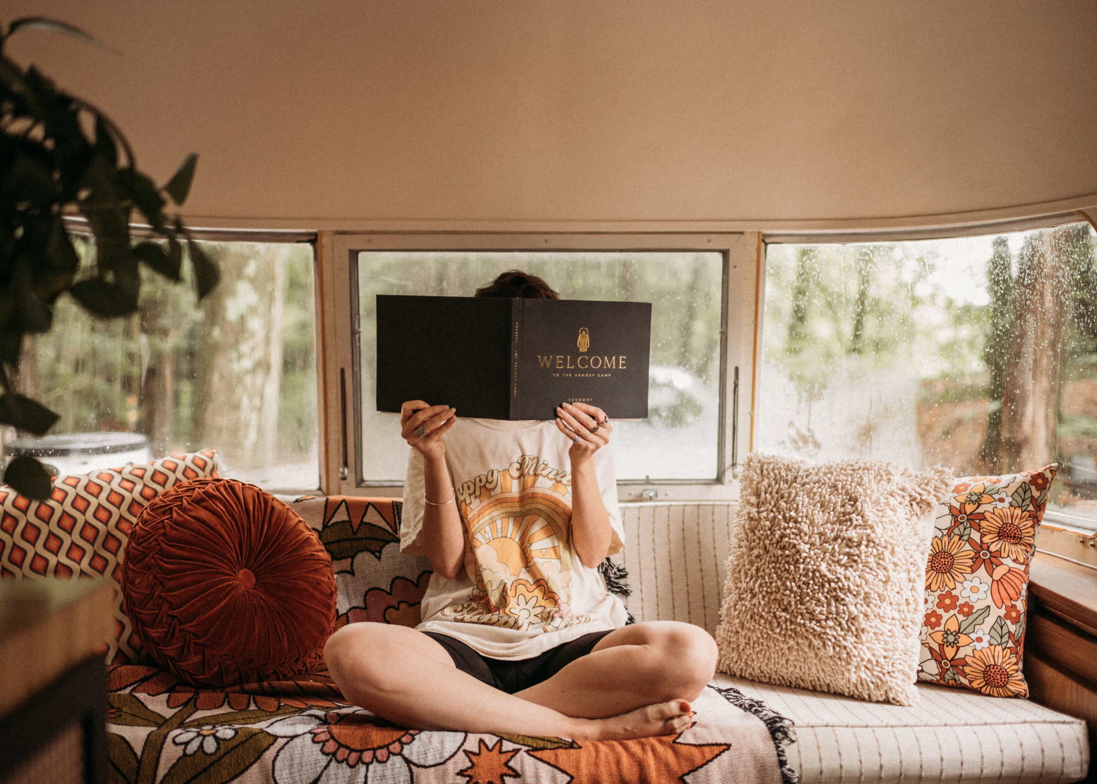 A person sitting cross-legged on a cushioned bench holds up a menu while surrounded by colorful cushions in a cozy, sunlit space.
