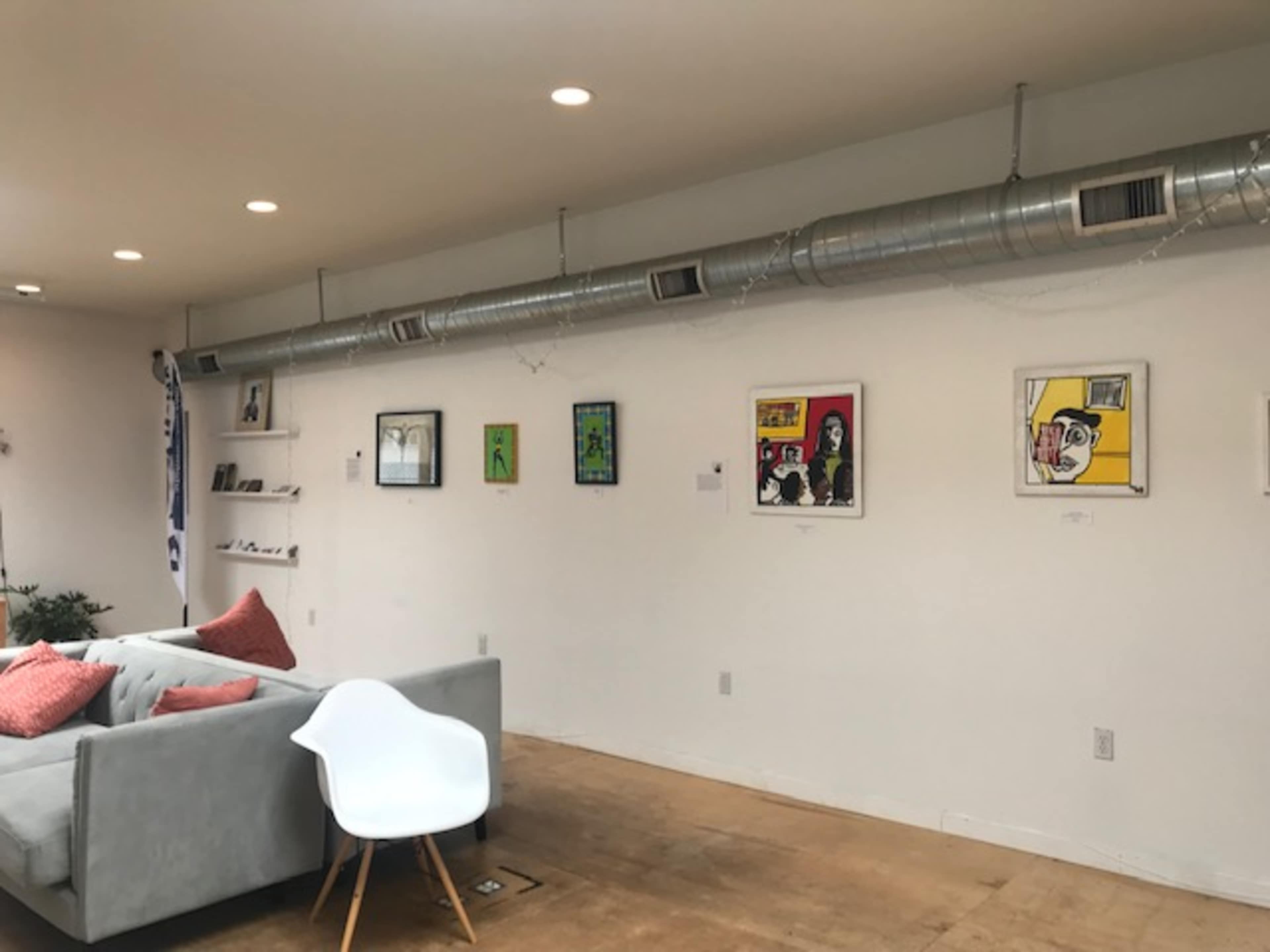 The image shows a modern room with a gray sofa, pink cushions, and a wall featuring framed artwork and exposed air ducts above.