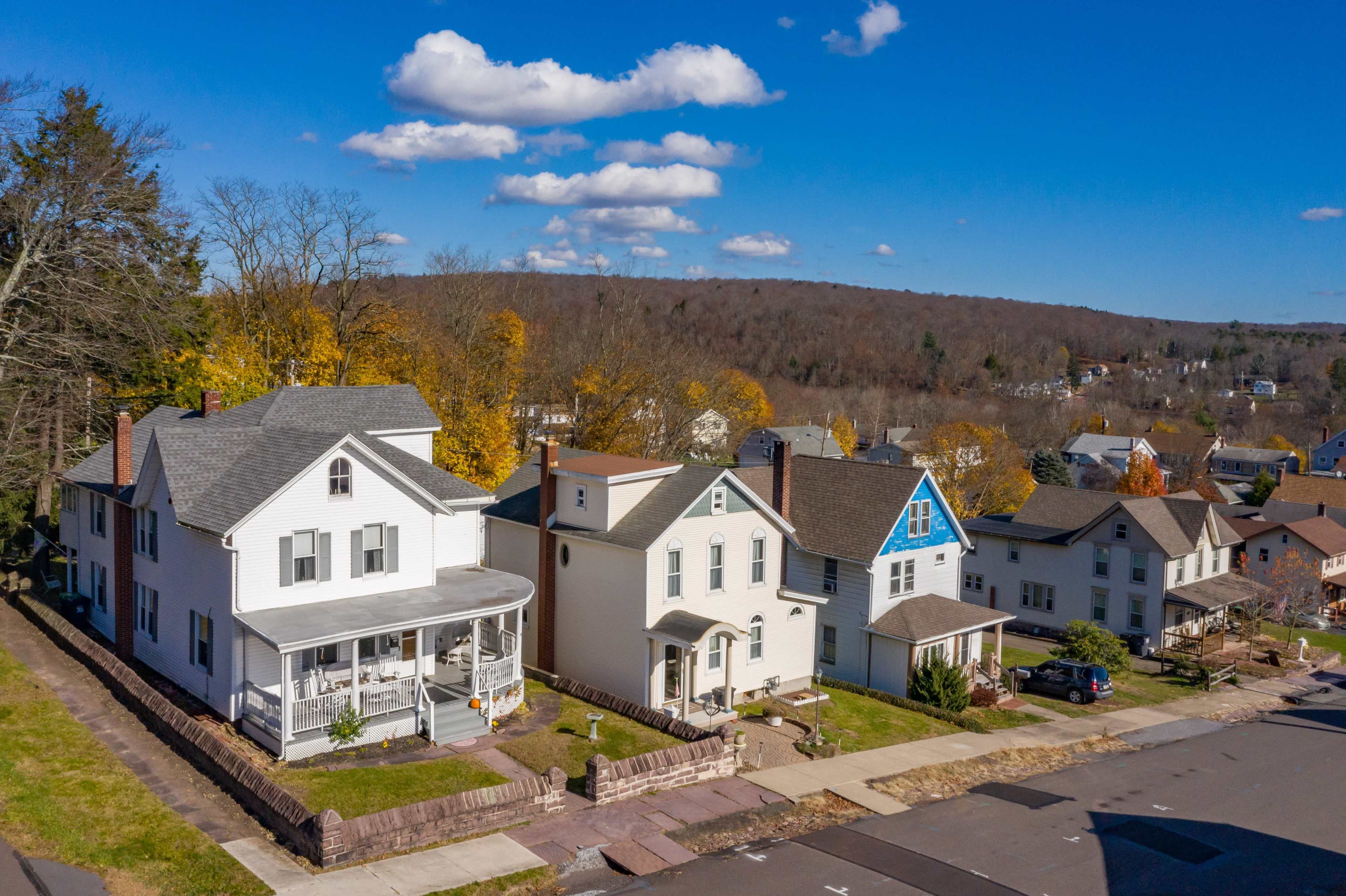 A row of residential houses with varied architectural styles and colors, set against a backdrop of trees and hills under a clear blue sky.