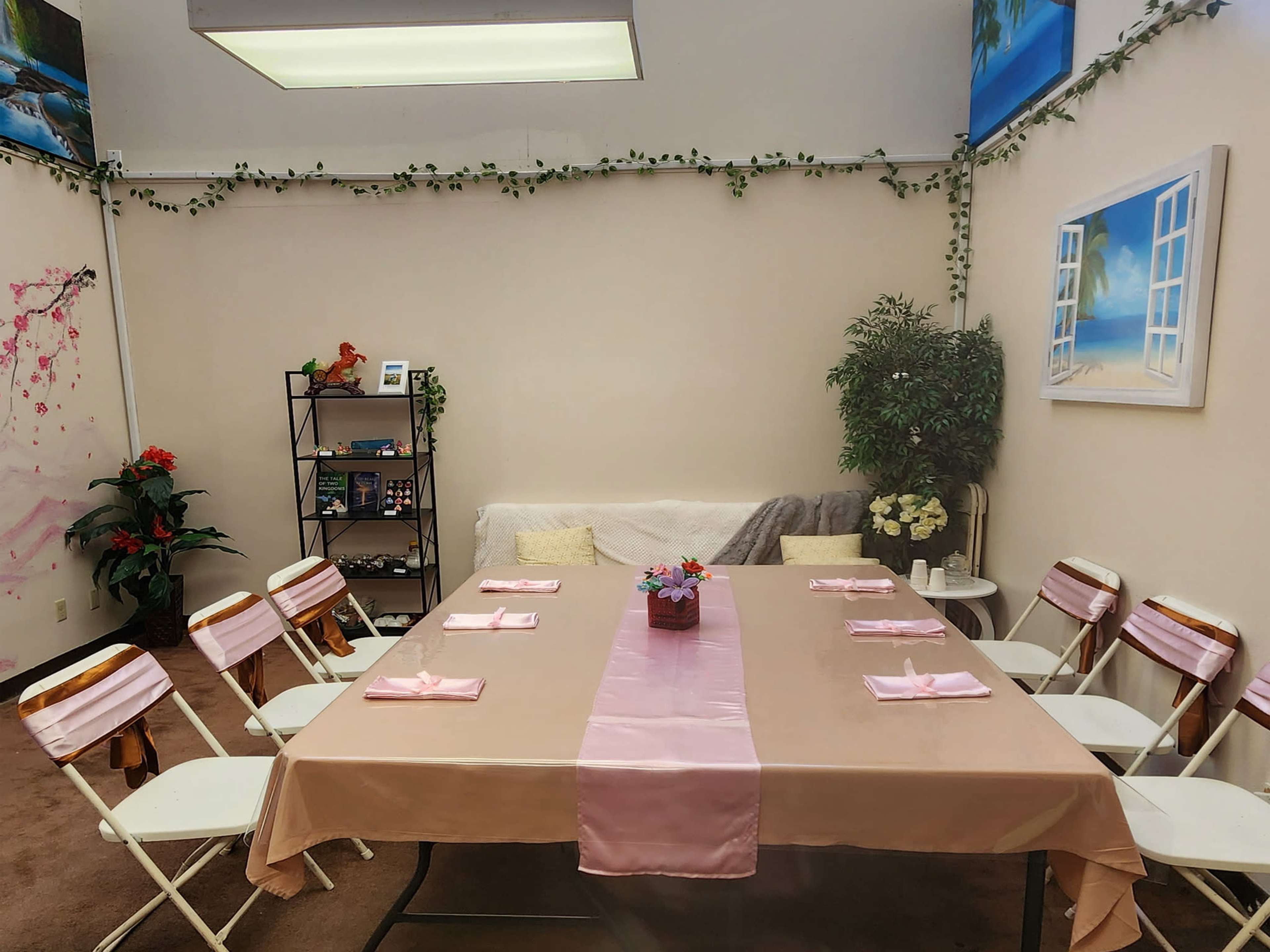 The image shows a decorated room with a long table set for a gathering, surrounded by eight chairs, with greenery on the walls and a few decorative items on shelves.