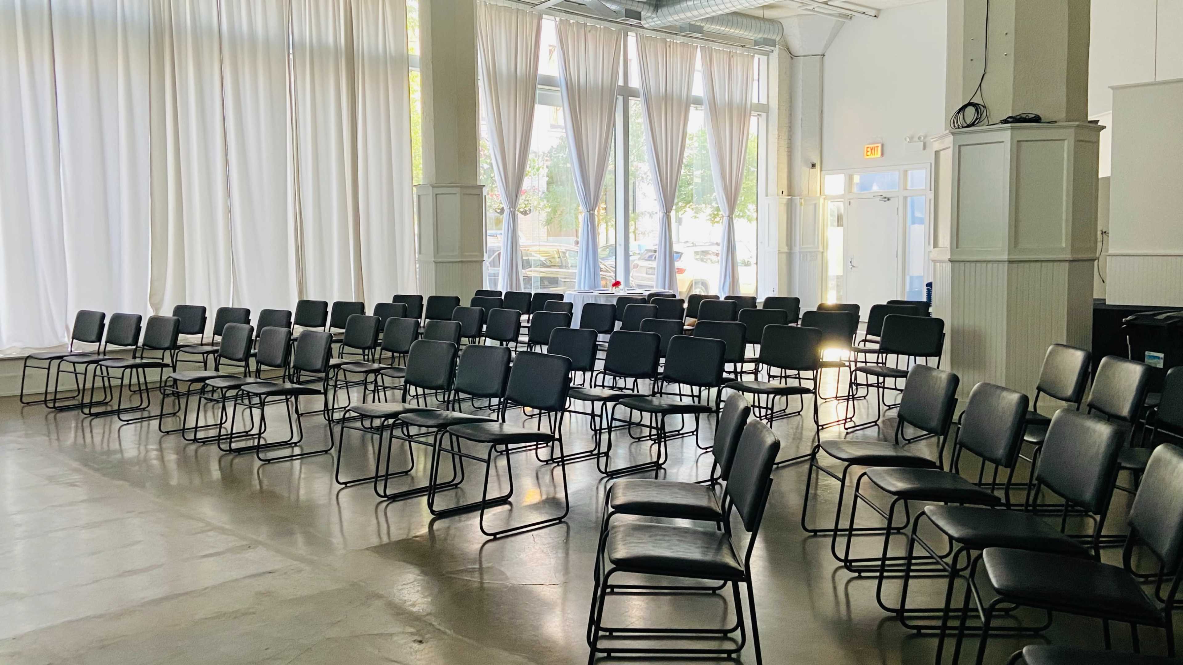 A row of black chairs is arranged in a spacious room with large windows and white curtains.