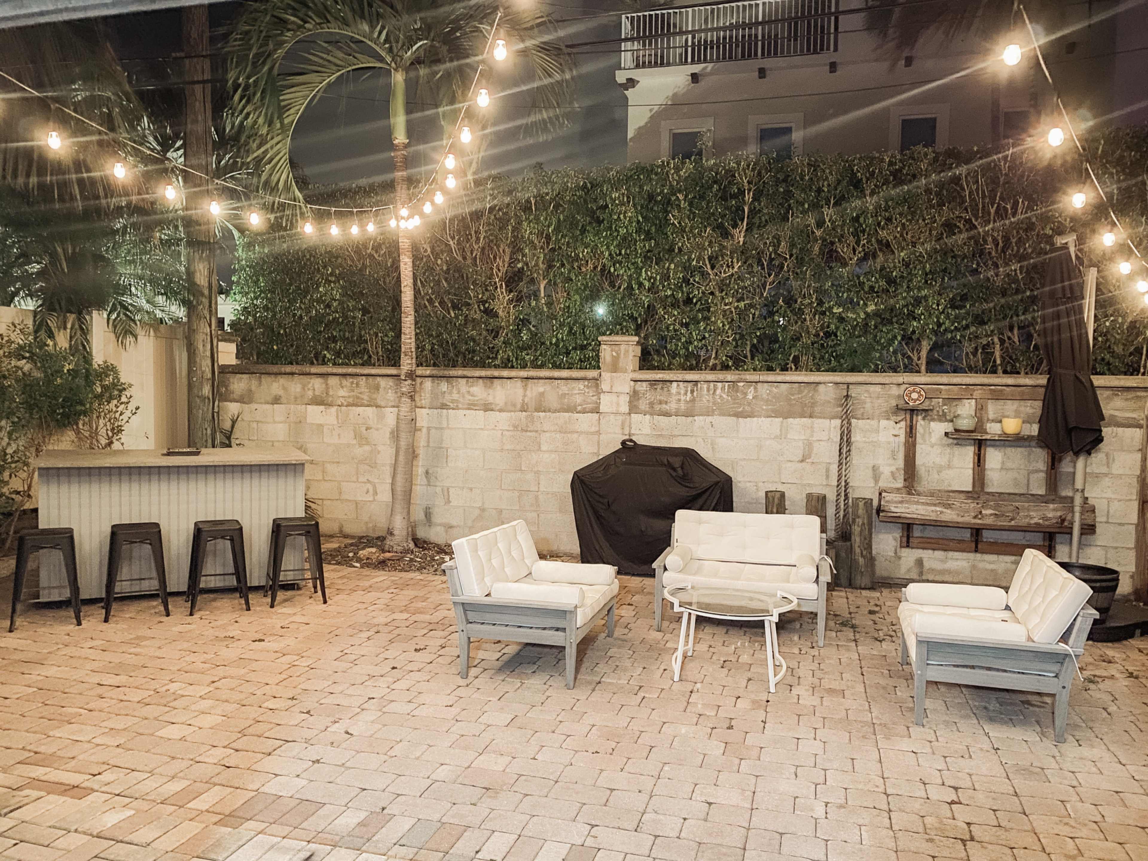 A patio area with four white chairs arranged around a small table, a bar counter with stools in the background, and string lights overhead.