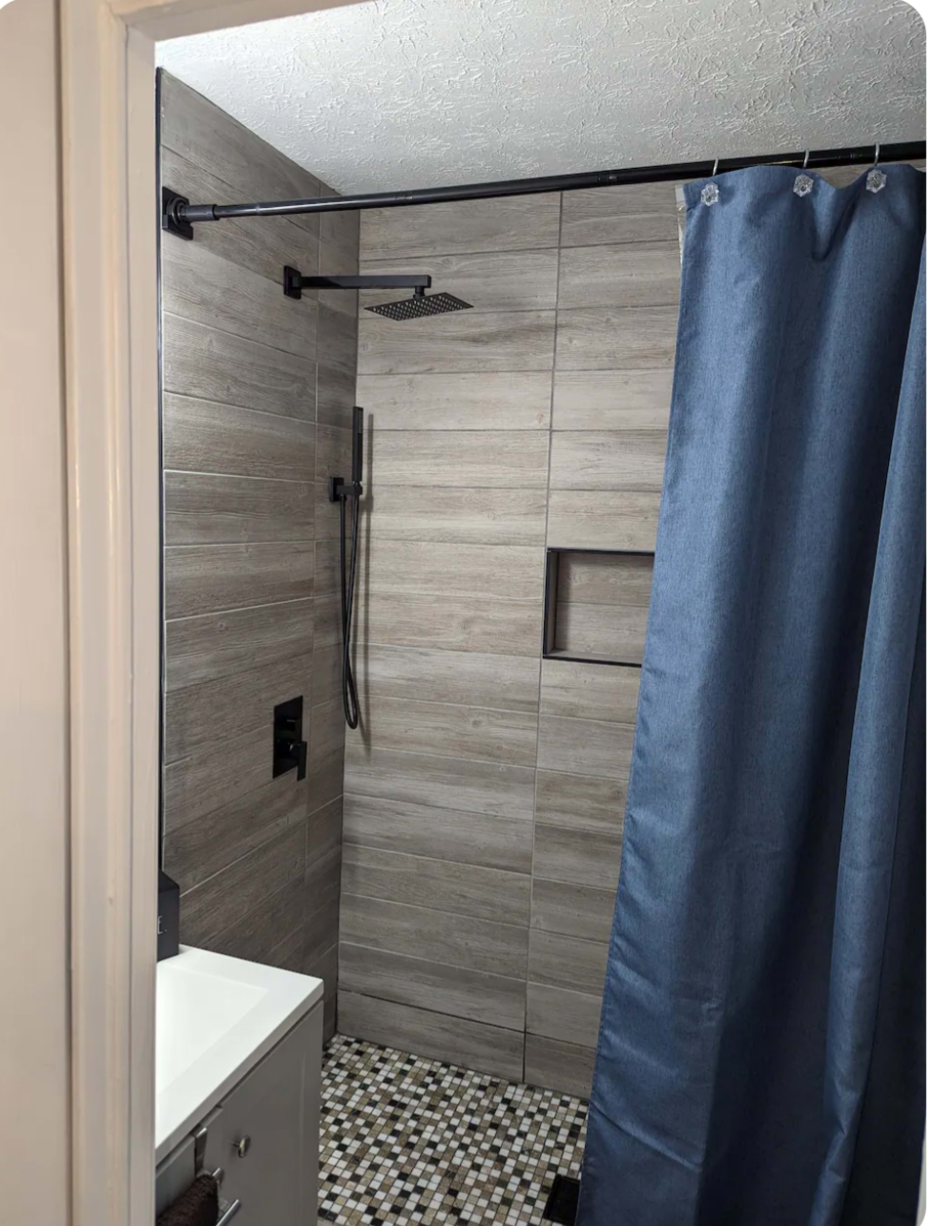 The image shows a modern shower space featuring beige tiled walls, a black shower fixture, and a blue shower curtain.