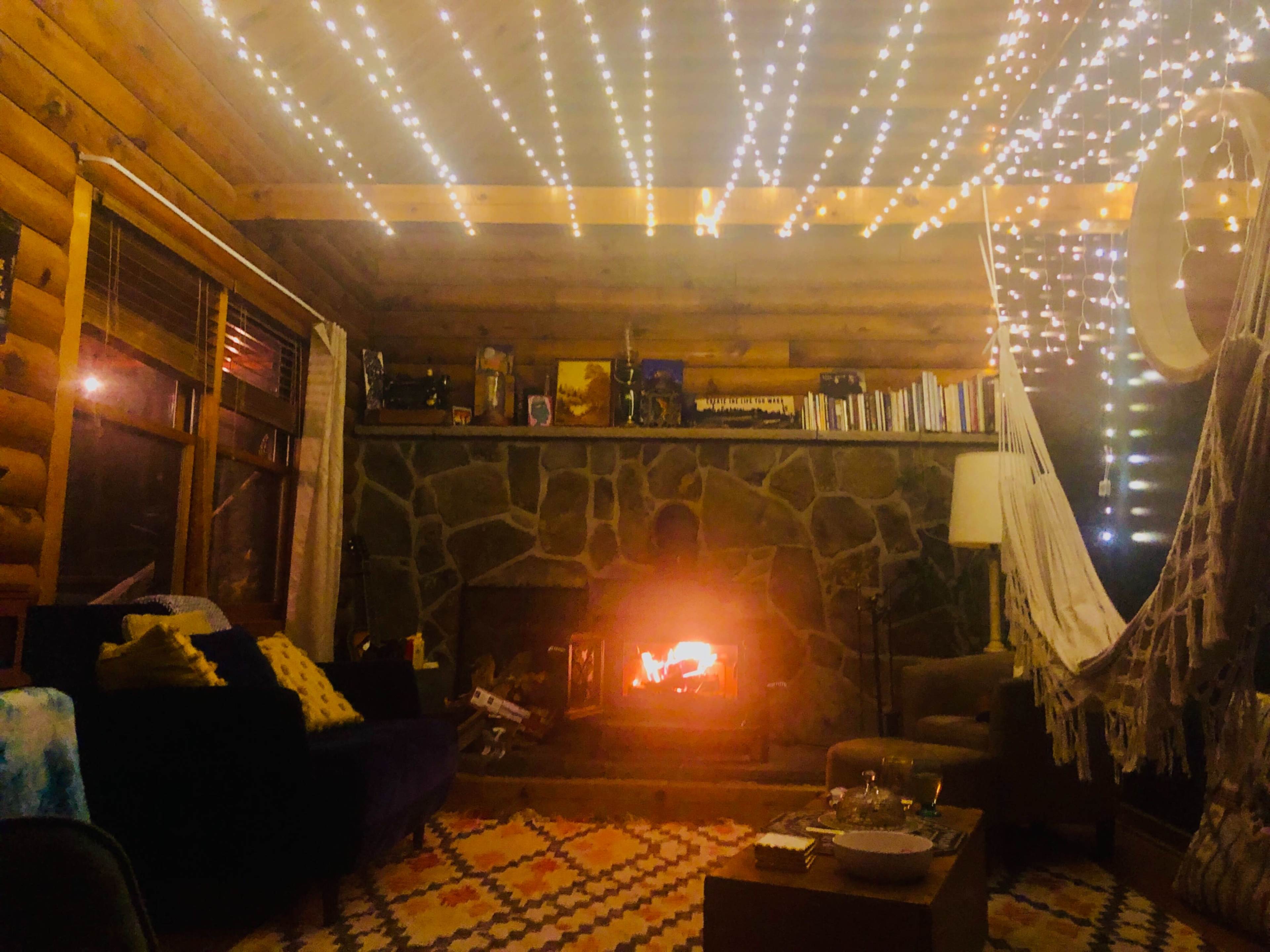 A cozy cabin interior features a stone fireplace with a glowing fire, surrounded by string lights and wooden walls.