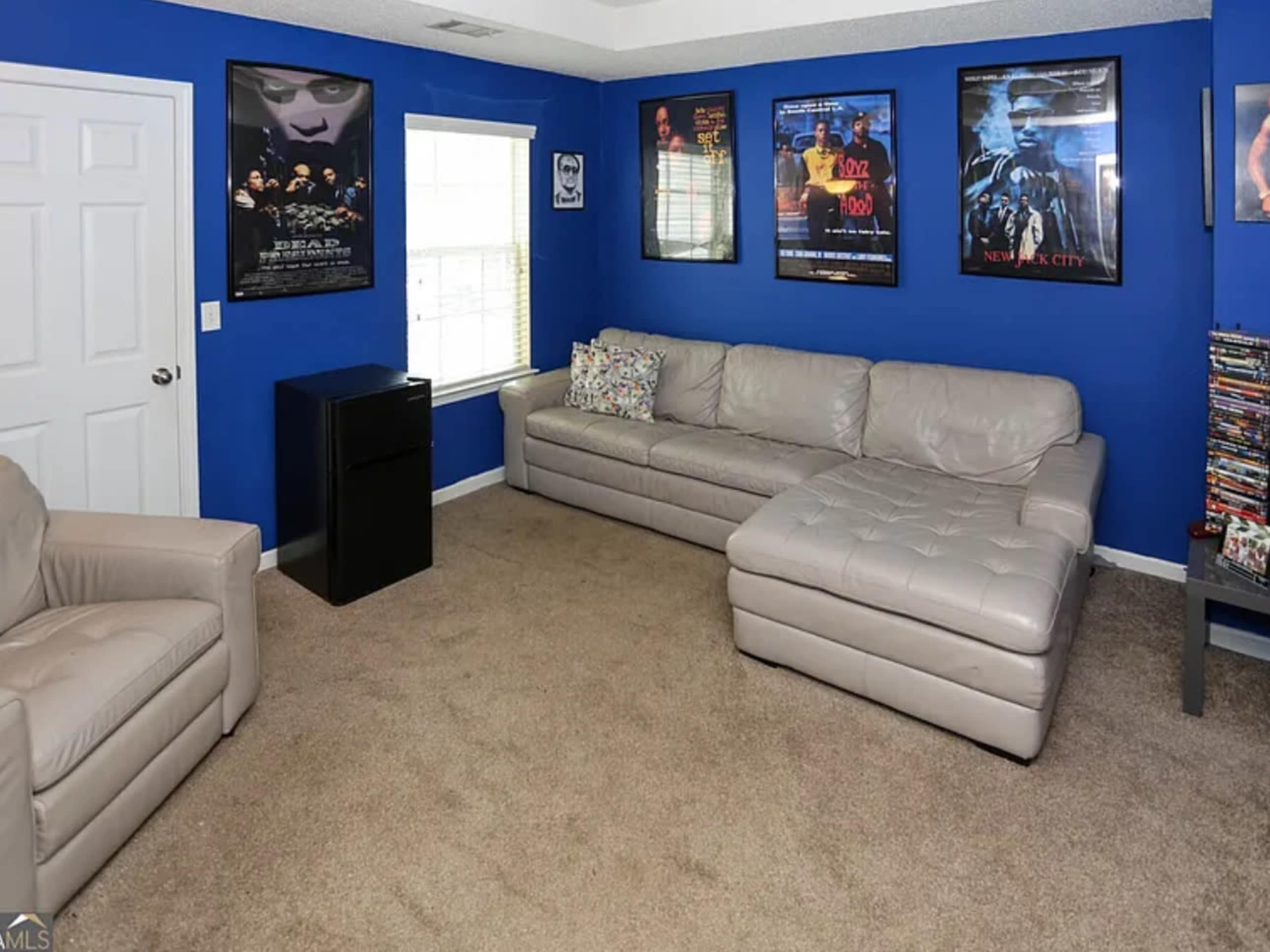 The room features a blue wall with several movie posters, a large beige sectional sofa, and a small black cabinet near a window.