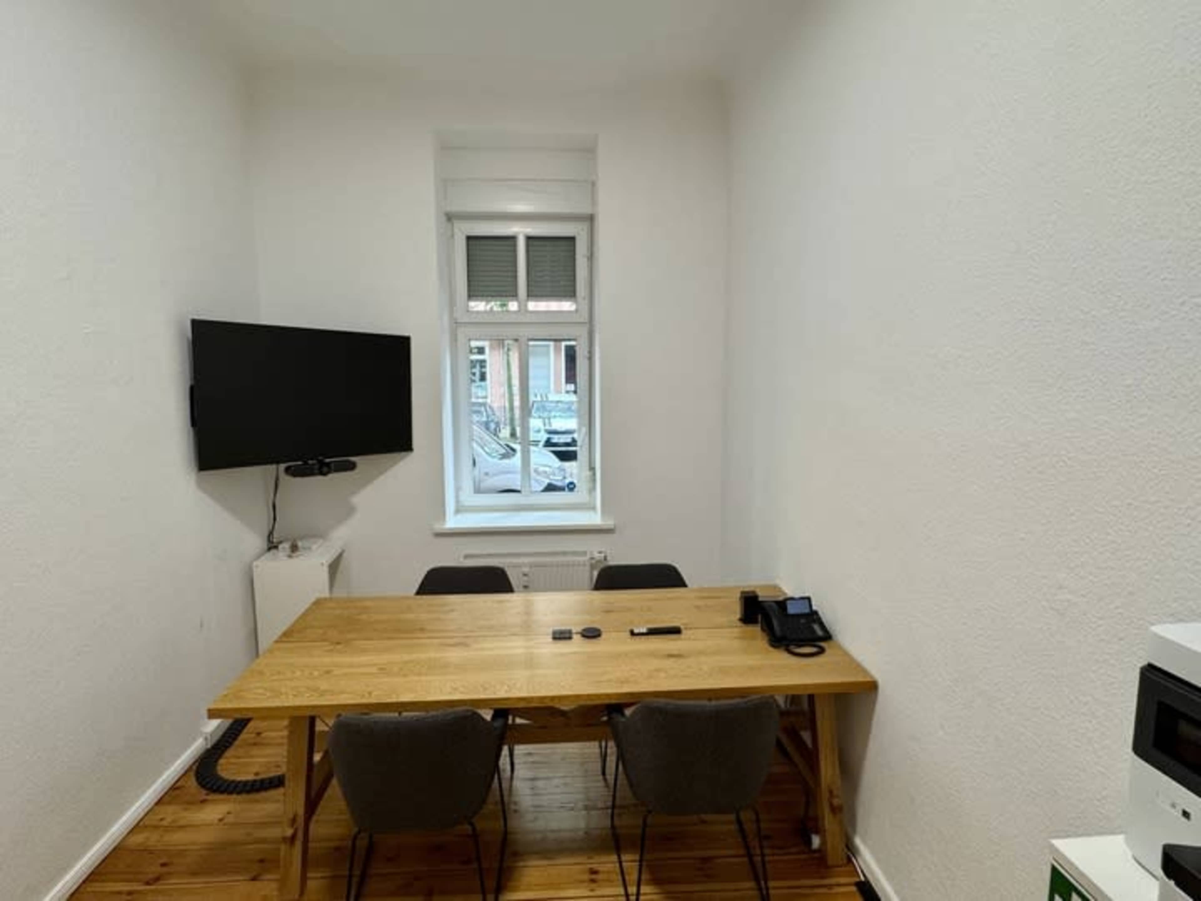The image shows a small conference room with a wooden table, four chairs, a wall-mounted TV, and a window looking out onto a street.