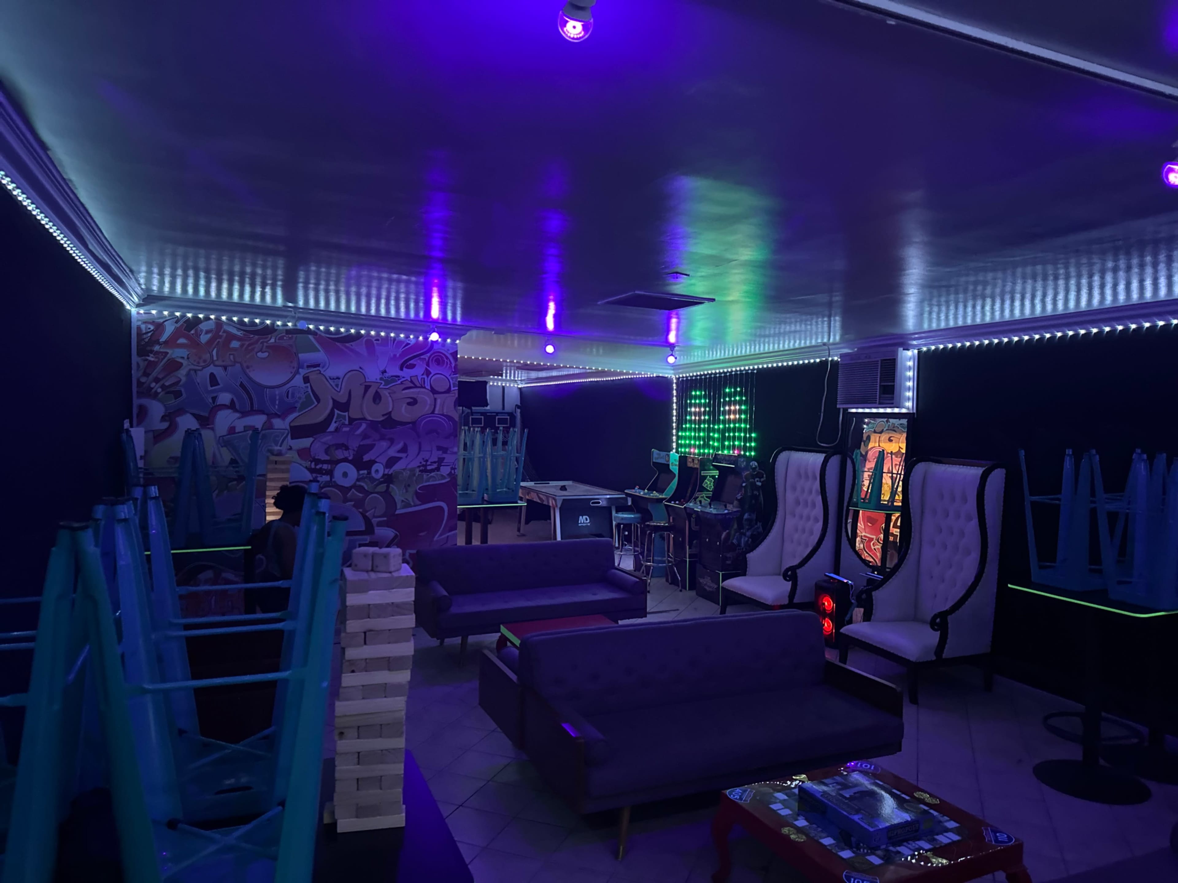 The image shows a dimly lit lounge area with colorful neon lights, empty chairs stacked upside down, and a Jenga tower on a table.
