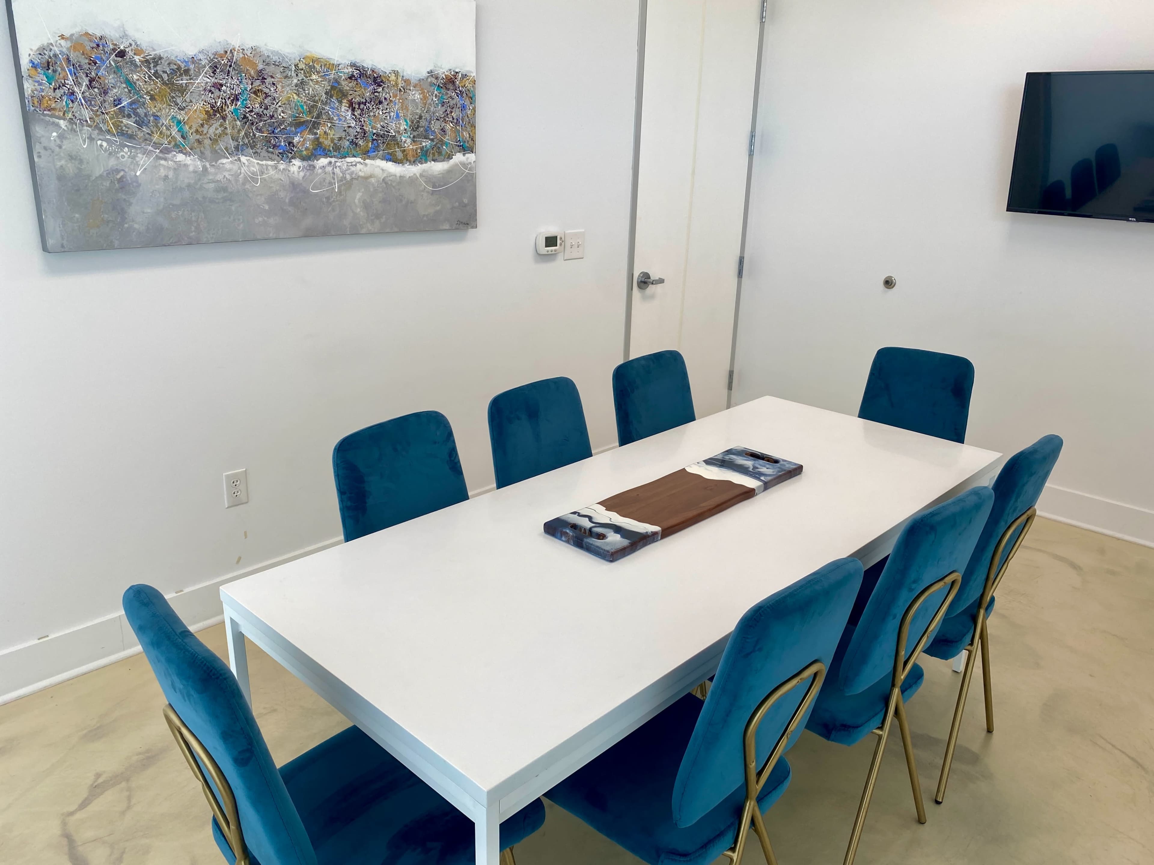 A modern conference room features a white table surrounded by eight teal chairs, with a piece of abstract art on the wall and a television mounted nearby.