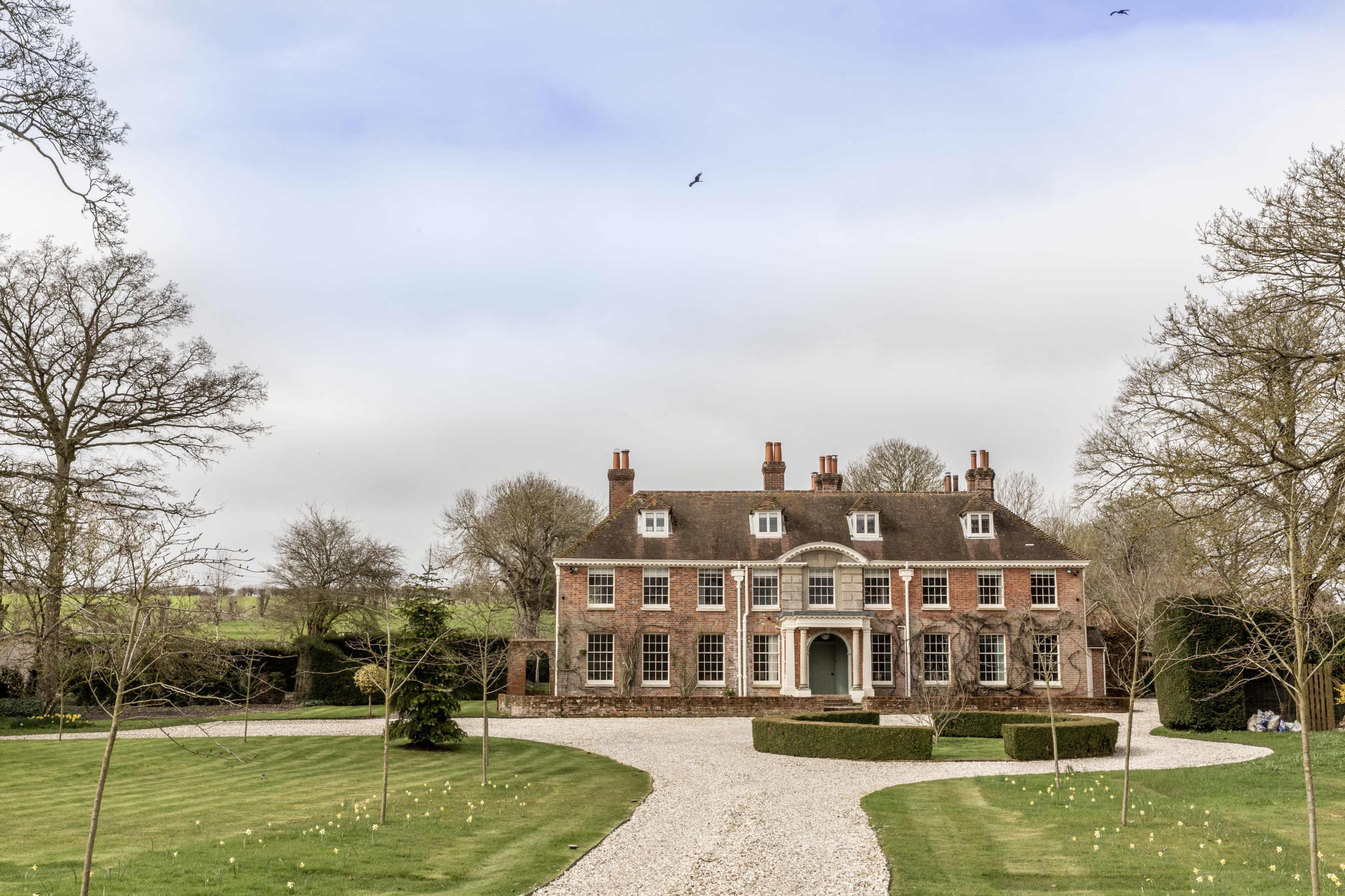 A large, brick mansion with a circular entrance is surrounded by a manicured lawn and gravel driveway.