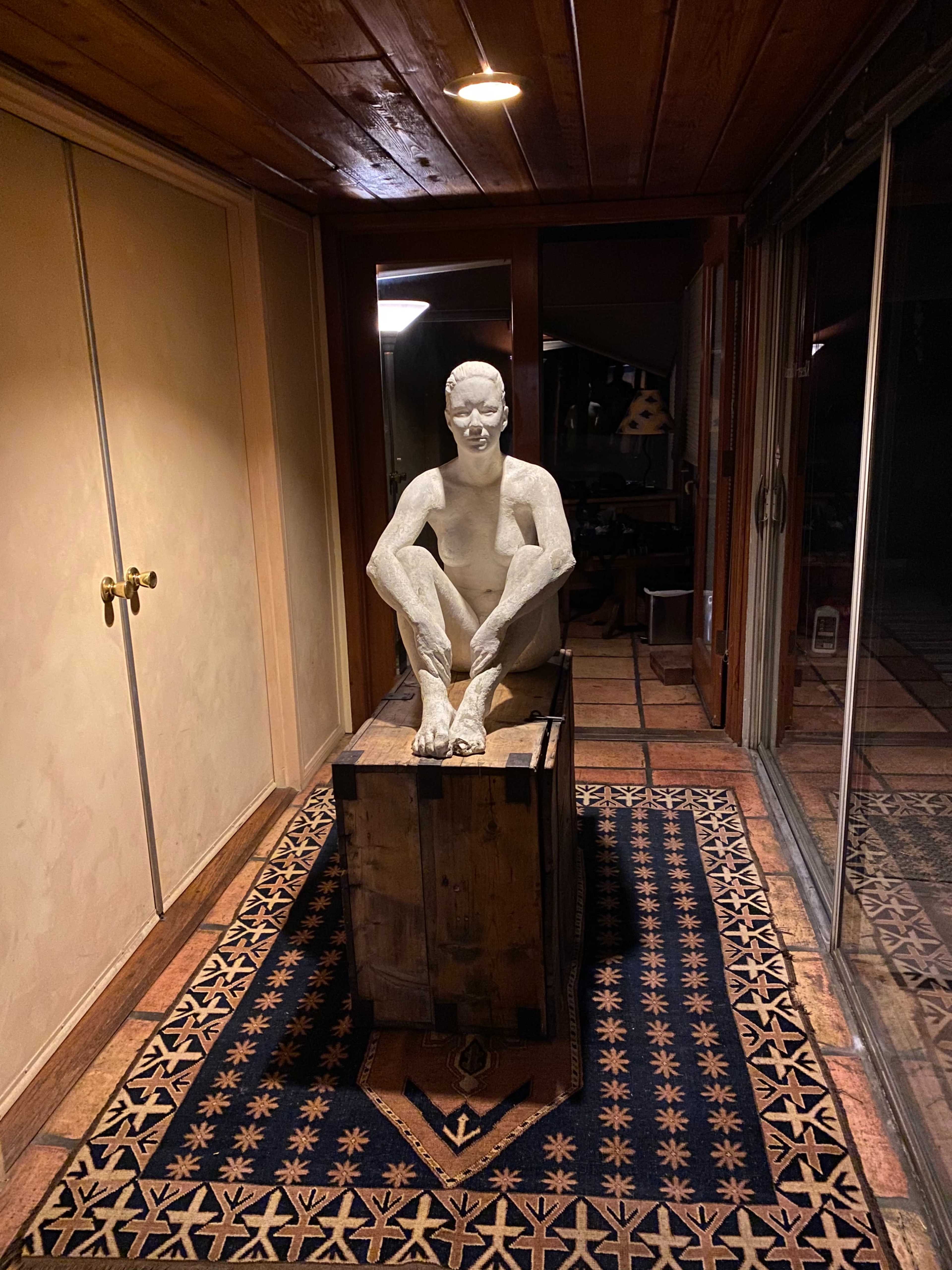 A seated sculpture made of a light-colored material sits atop a wooden box in a corridor, surrounded by tiled flooring and an ornate rug.