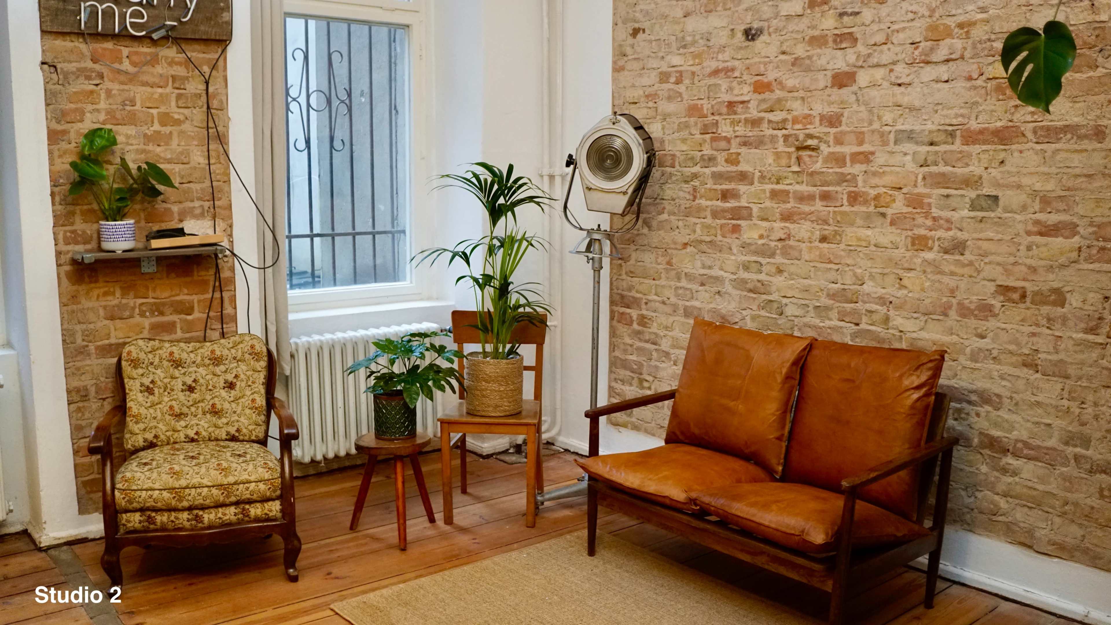 The image shows a cozy interior featuring a brick wall, two seating areas—one with a patterned chair and the other with a brown sofa—along with potted plants and a decorative fan.